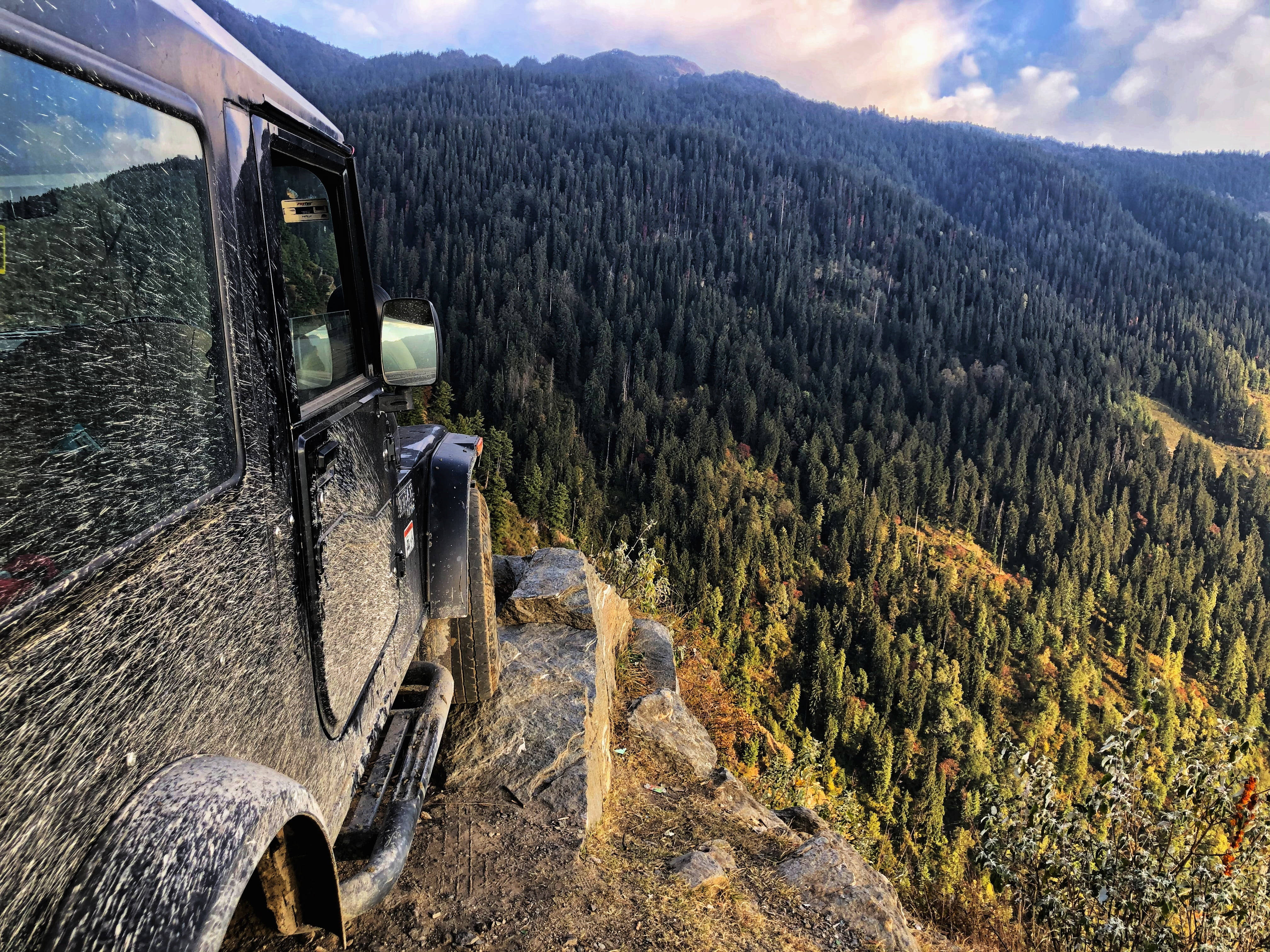 山景 桌布 壁紙 — 一輛泥濘的越野車棲息在懸崖邊緣，俯瞰著廣闊的森林山谷，沐浴在金色的日落光芒中。