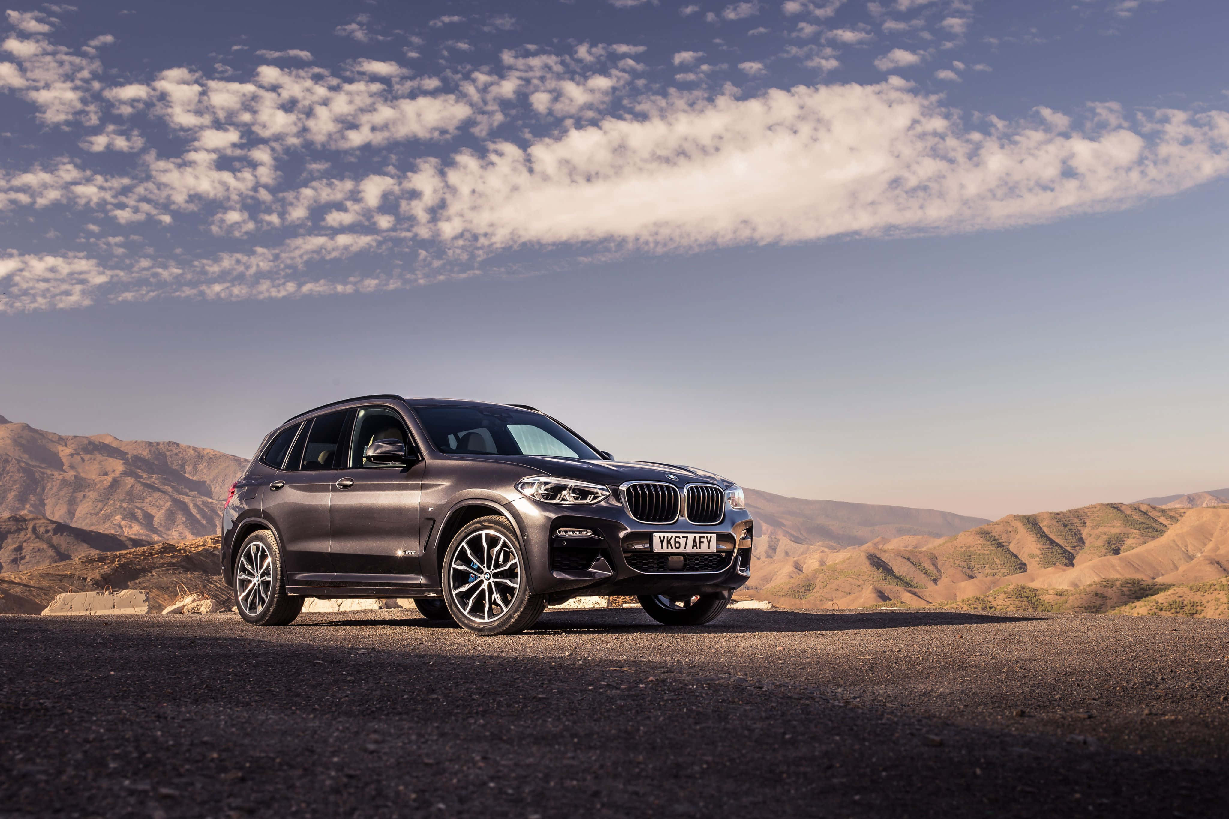 Bmw X3 桌布 壁紙 — 一輛流線型啞光灰色BMW X5 SUV停泊在沙漠公路上，背景是起伏的棕色山丘和黃金時段部分多雲的天空。