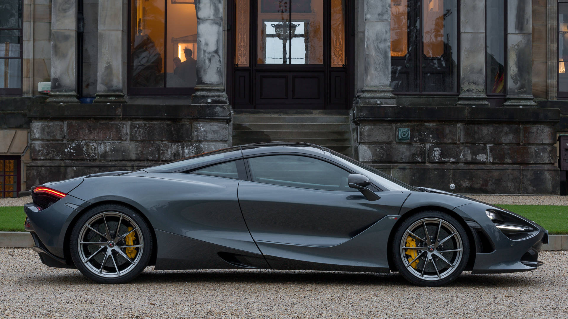 mclaren 720s 4K 桌布 壁紙 — 一輛流線型啞光灰色McLaren 720S跑車停泊在碎石上，前面是一座有溫暖室內燈光的大型石建築。