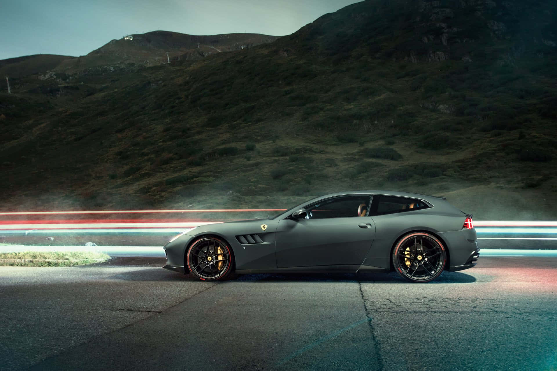 Ferrari Gtc4lusso 桌布 壁紙 — 一輛流線型啞光灰色法拉利跑車在黃昏時分沿著山路行駛，伴隨著過往車輛留下的光跡。