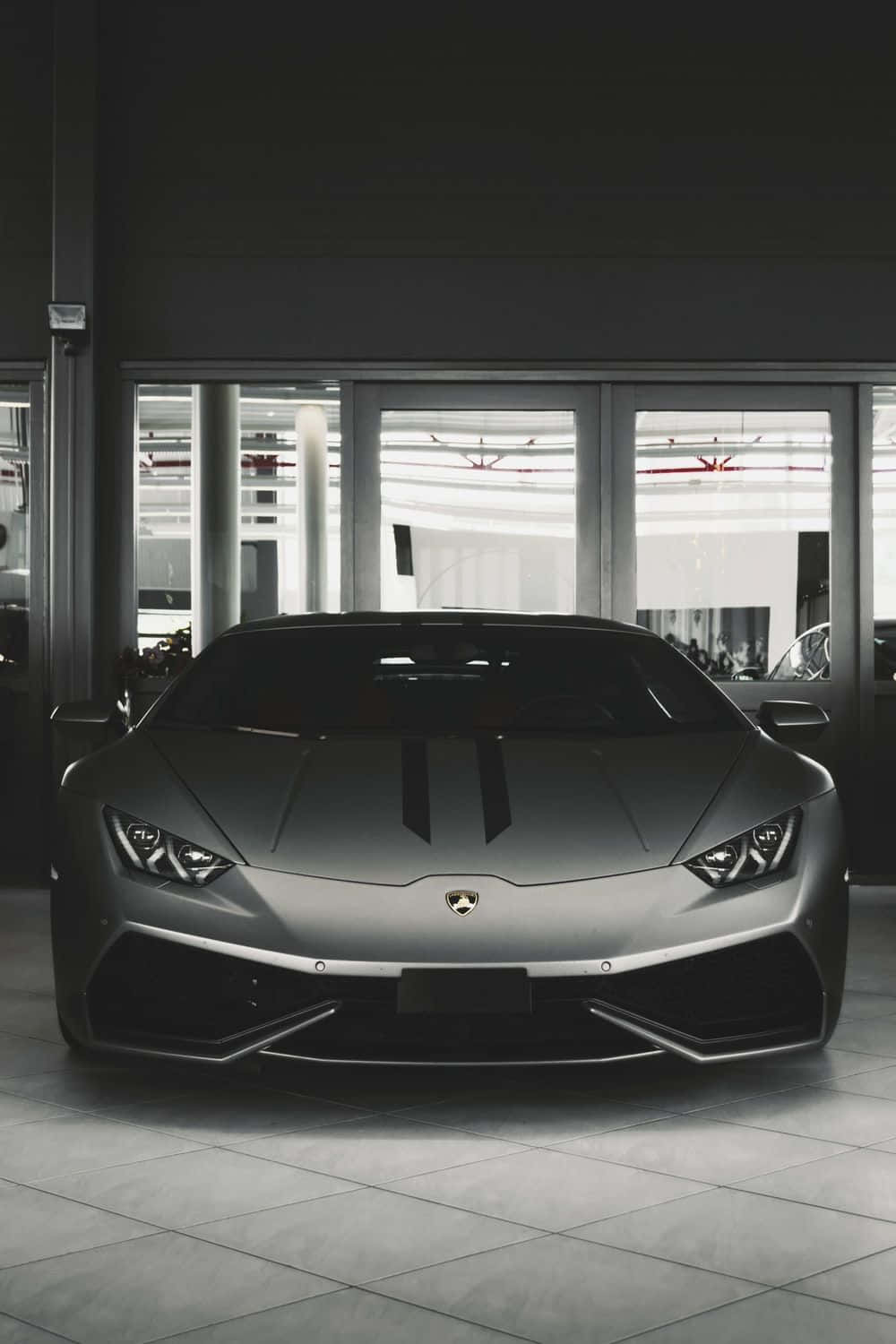 Black And White Car 桌布 壁紙 — 一輛流線型啞光灰色藍寶堅尼跑車帶有黑色賽車條紋，位於現代簡約的展廳中央，地板鋪有瓷磚，窗戶寬大。