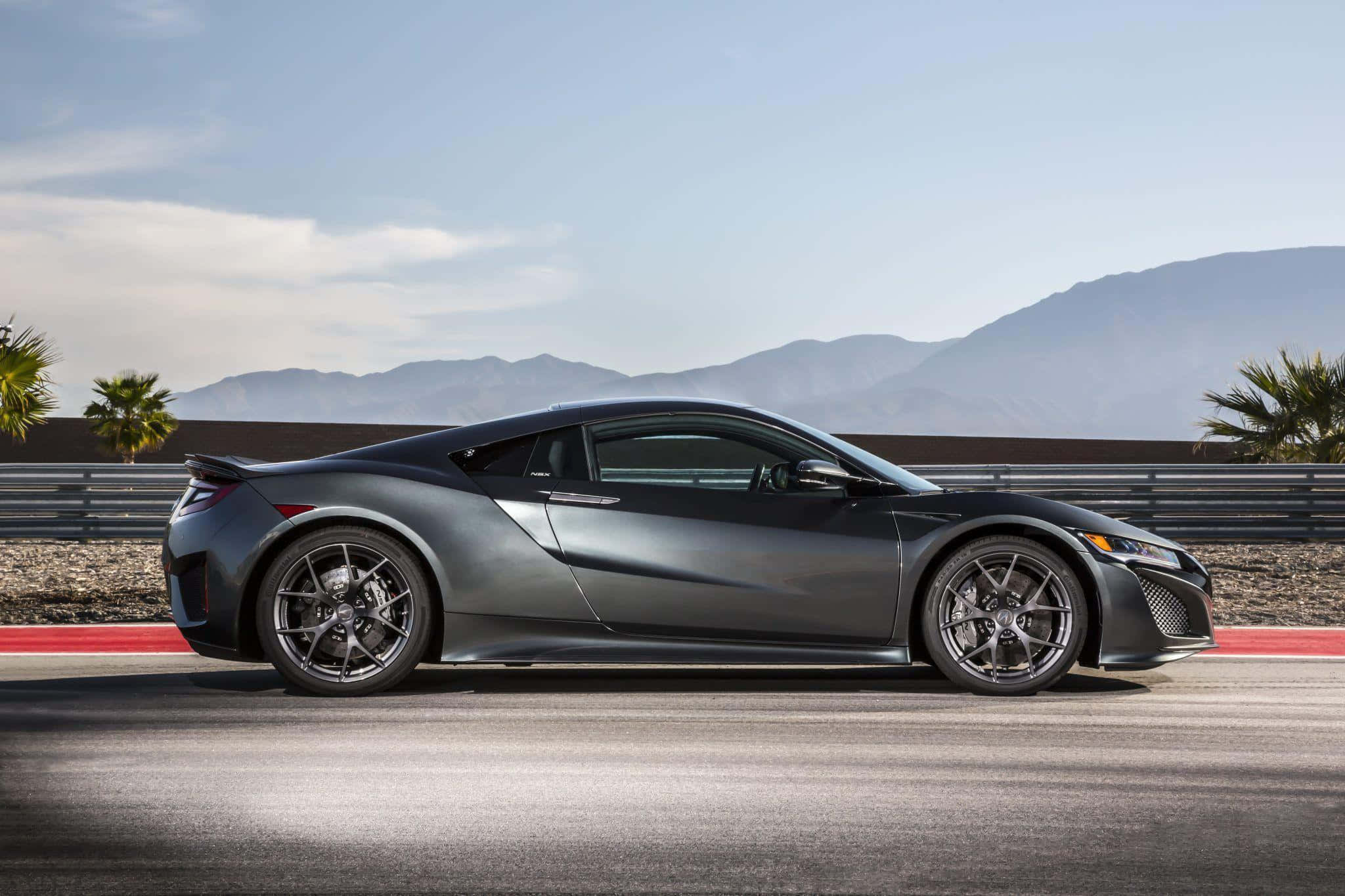 Acura Nsx 桌布 壁紙 — 一輛流線型啞灰色跑車在晴朗天空下，於有遠山和棕櫚樹的沙漠賽道上行駛。
