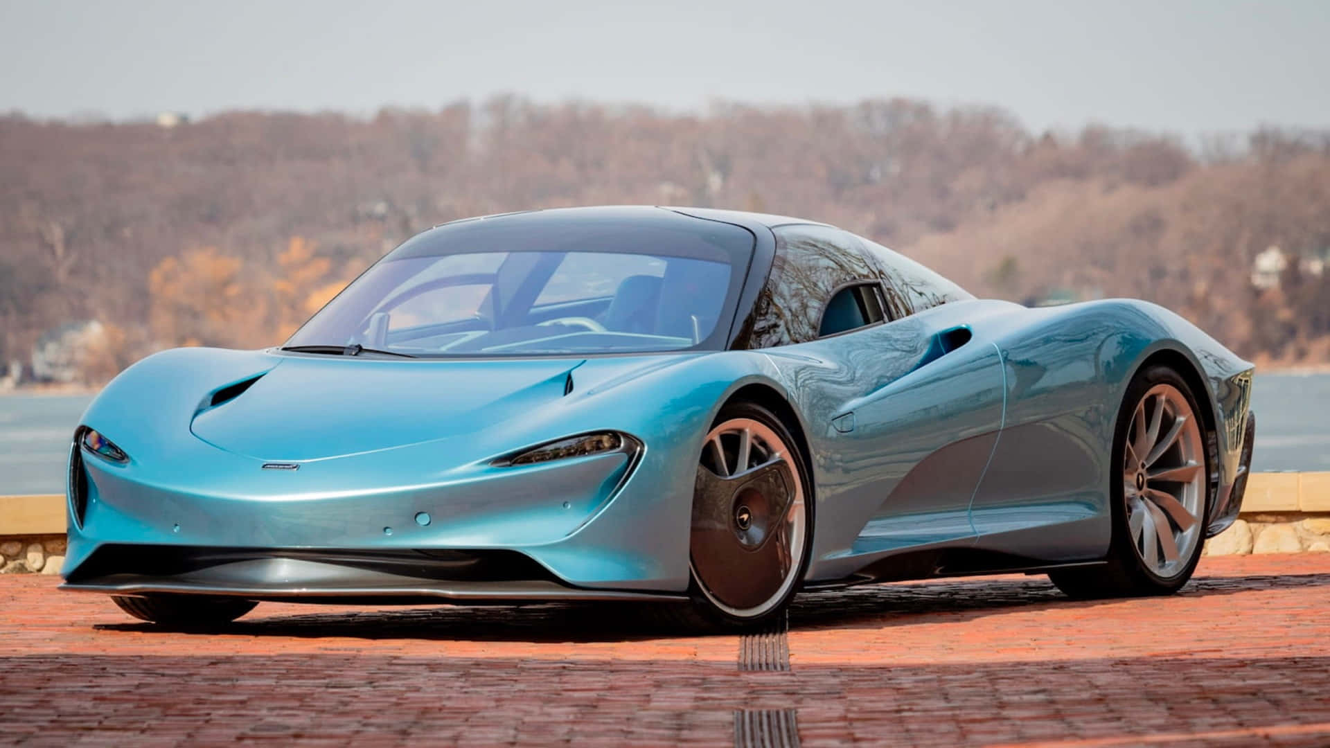 Mclaren Speedtail 桌布 壁紙 — 一輛流線型淺藍色跑車，帶有空氣動力學曲線，停在紅磚路上，旁邊是水，背景有光禿禿的樹木。