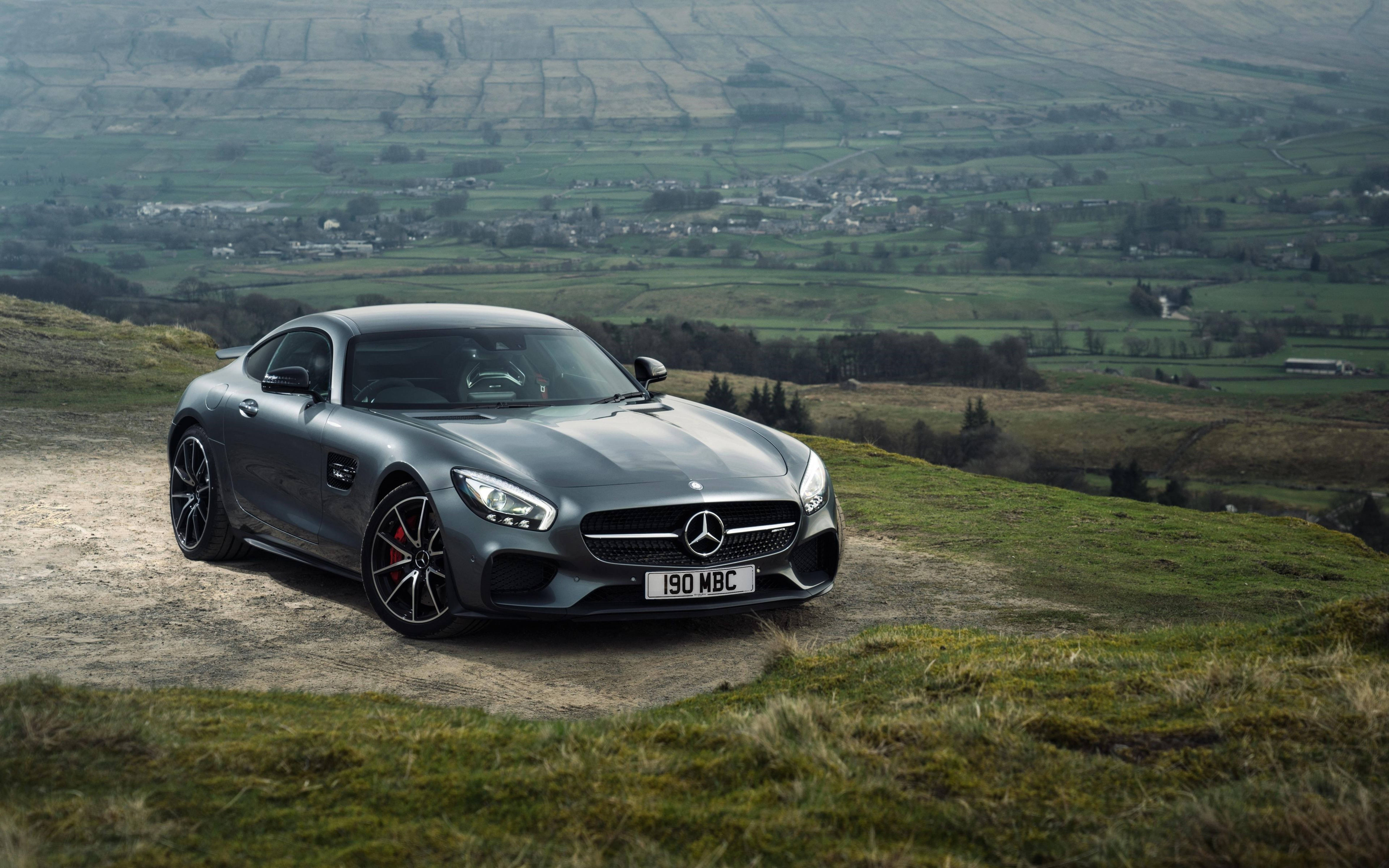 跑車 桌布 壁紙 — 一輛流線型灰色 Mercedes-AMG GT 跑車停泊在草坡上，俯瞰著一個廣闊的綠色山谷，山谷中散落著房屋。