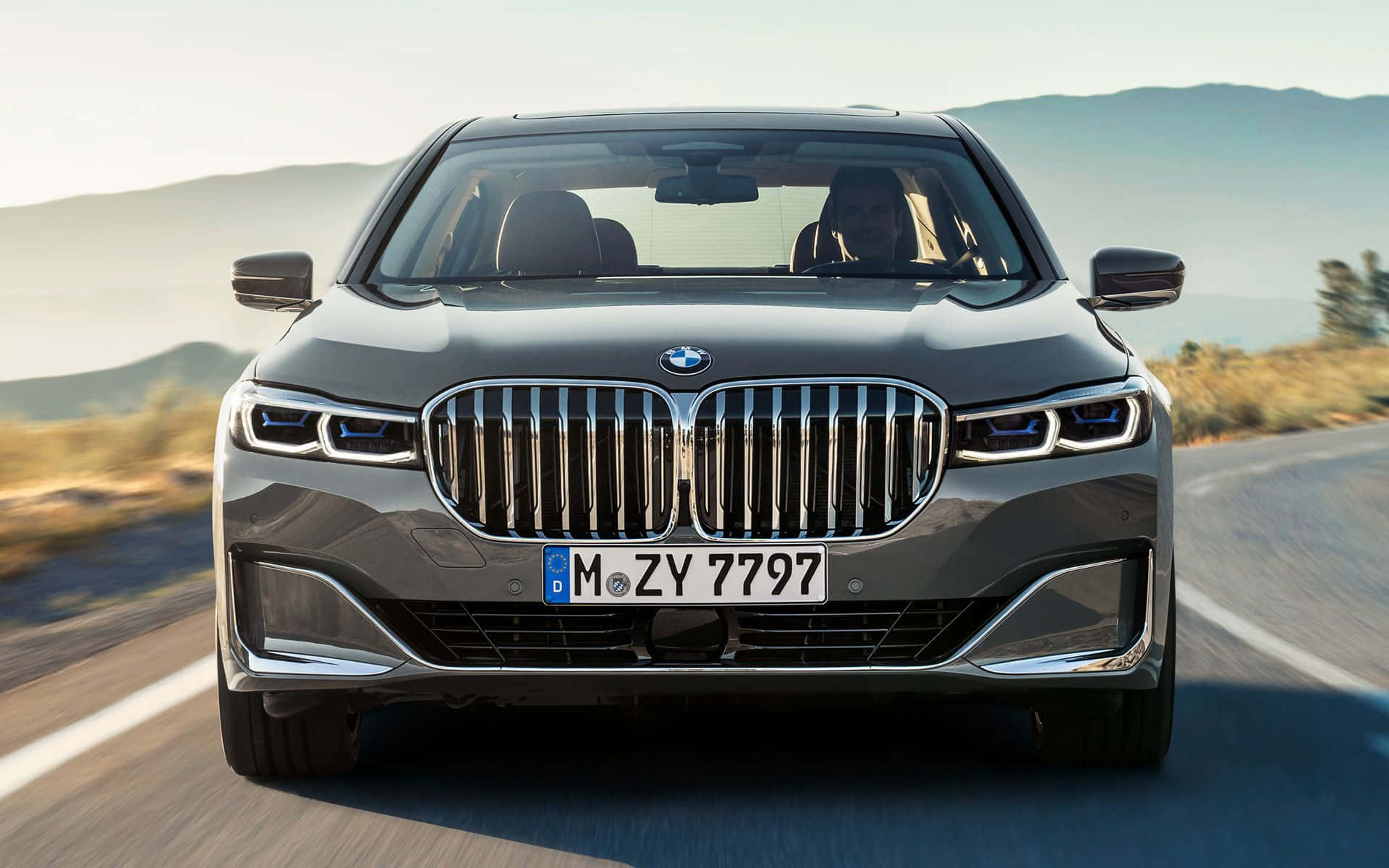 Bmw 7 Series 桌布 壁紙 — 一輛流線型灰色BMW 7系列轎車在陽光明媚的山路上向前行駛，背景模糊。