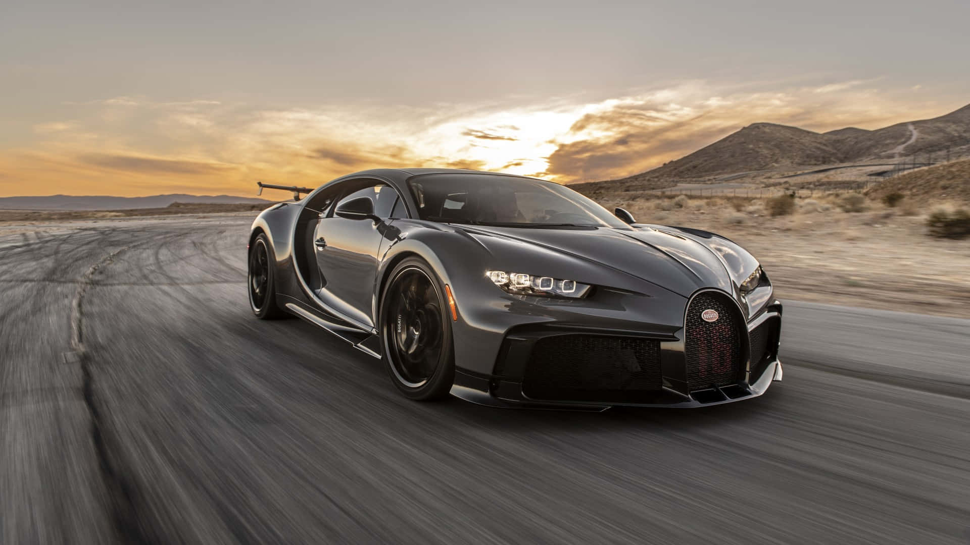 1920 X 1080 Car 桌布 壁紙 — 一輛流線型灰色Bugatti Chiron在日落時分沿沙漠公路飛馳，帶有模糊動態和遠山。
