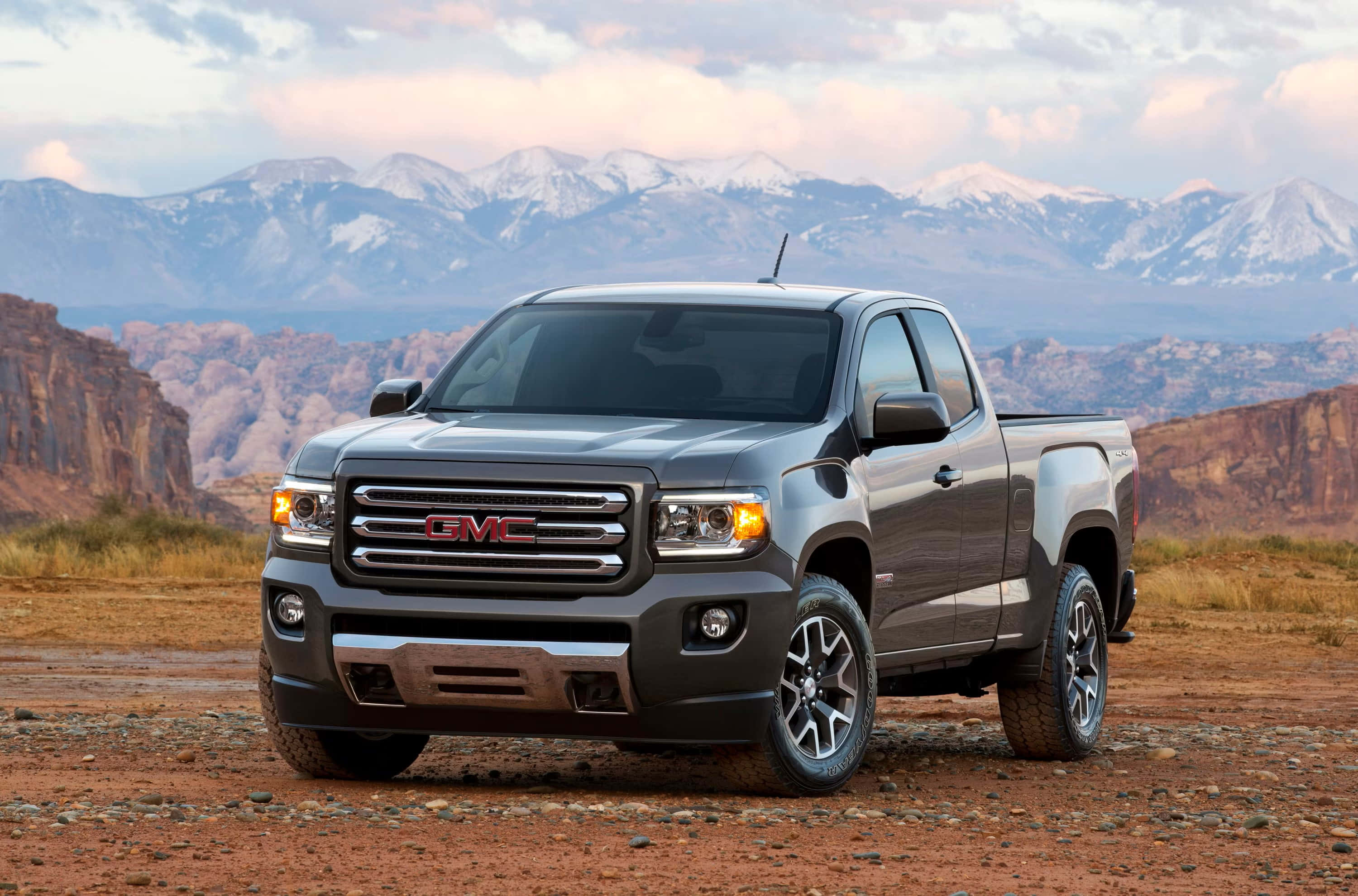Gmc Canyon 桌布 壁紙 — 一輛流線型灰色GMC Sierra皮卡車停在崎嶇的沙漠小徑上，背景中白雪覆蓋的山脈在柔和暮色下巍峨聳立。