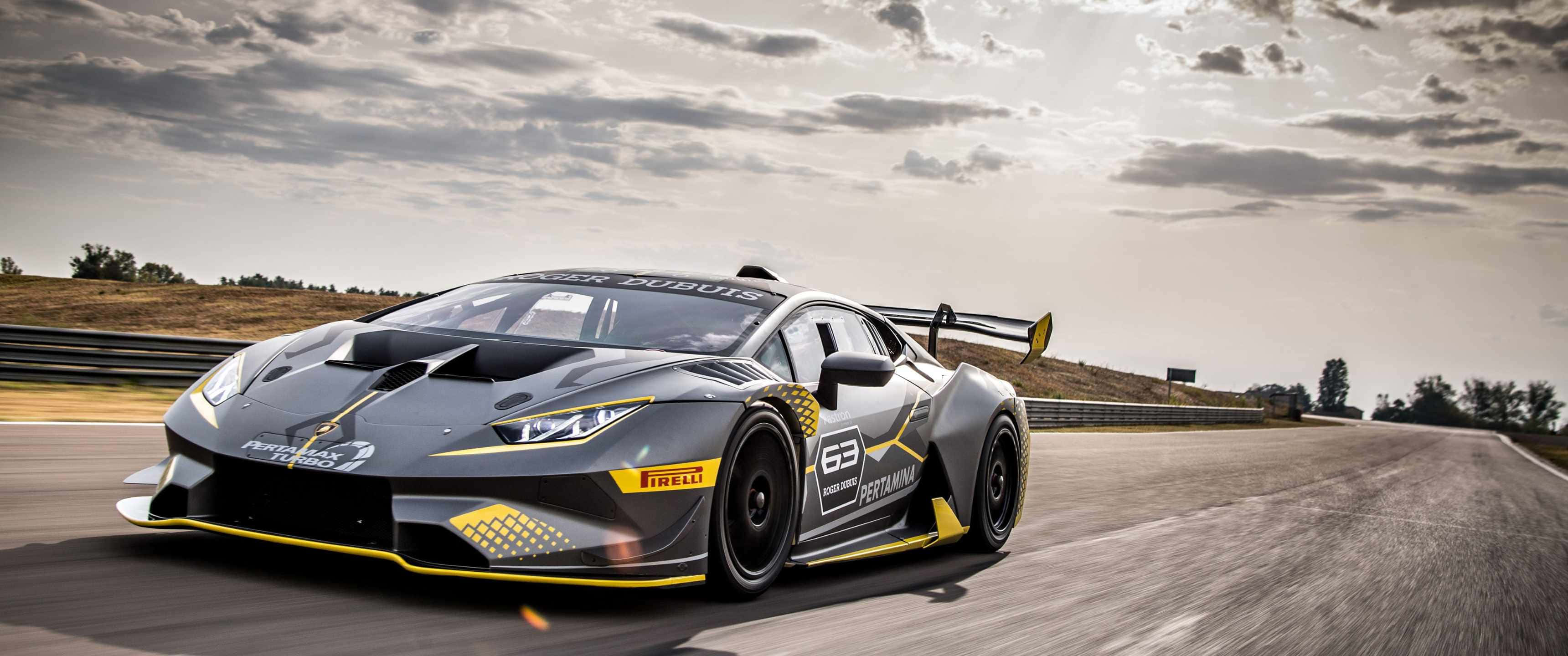 3440x1440p Lamborghini 桌布 壁紙 — 一輛流線型灰色和黃色藍寶堅尼賽車在陽光普照的賽道上奔馳，天空多雲。