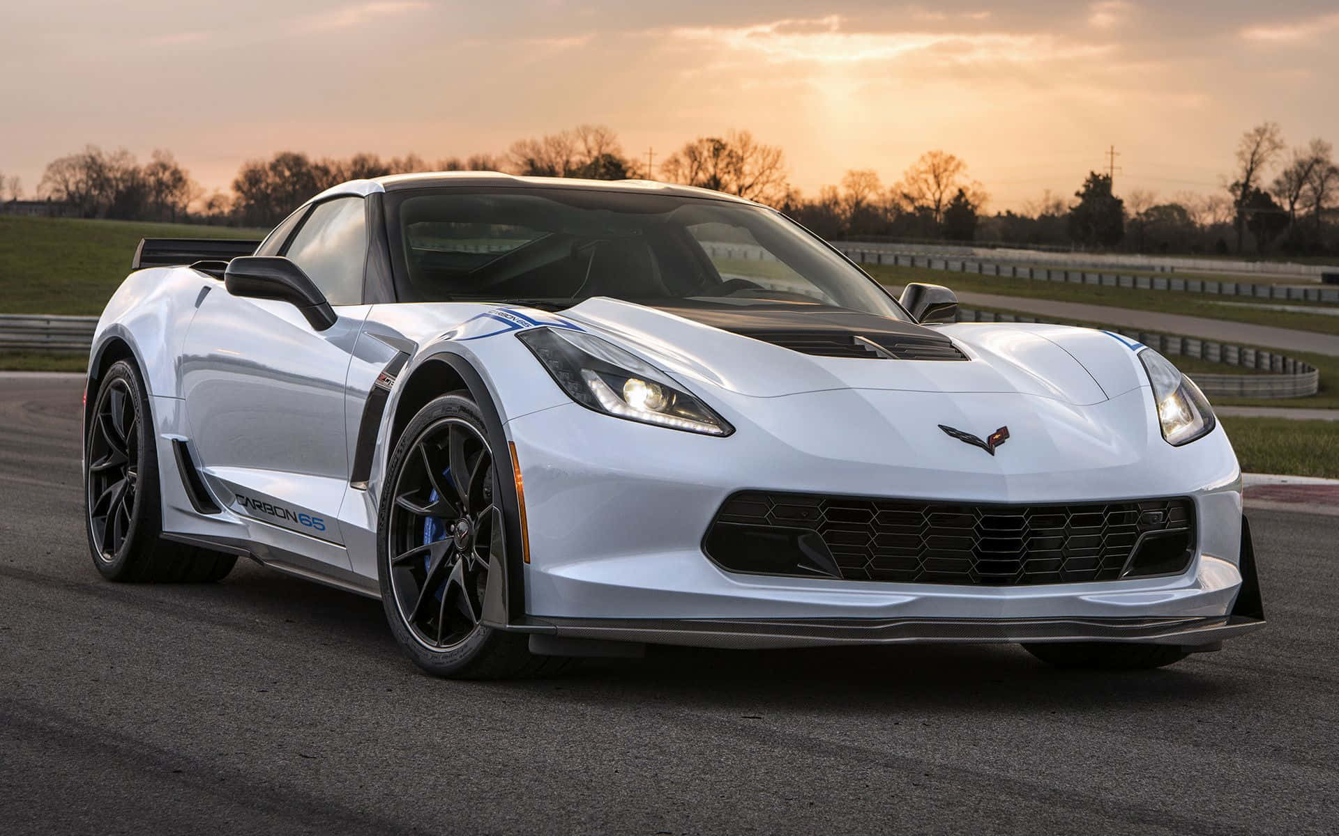 Chevrolet Corvette Z06 桌布 壁紙 — 一輛流線型白色 Corvette C7.R 跑車在金色夕陽下、背景有光禿樹木的賽道上閃閃發光。