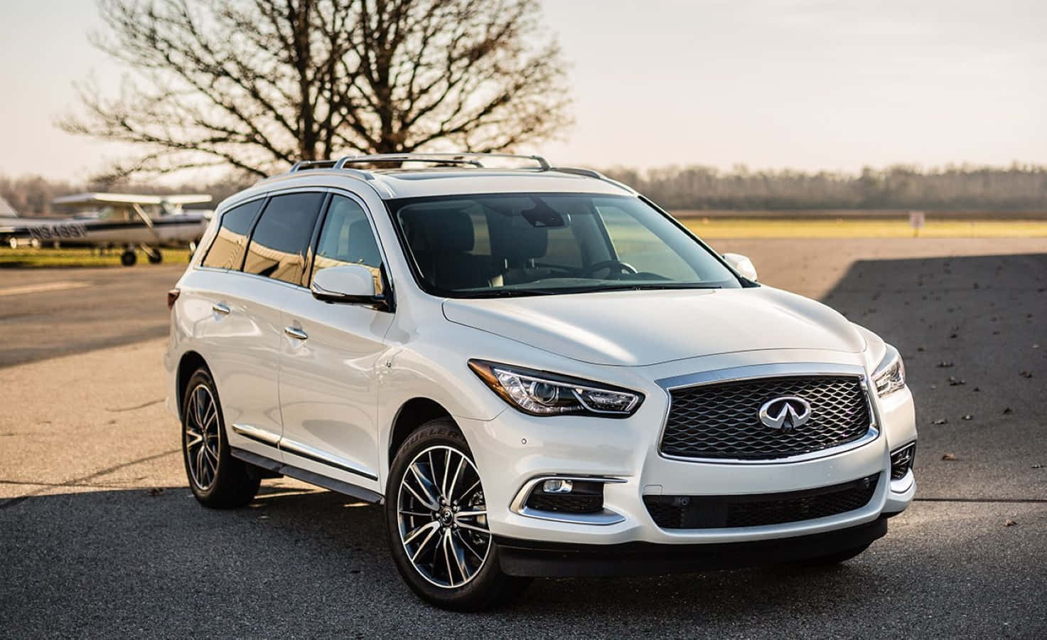 Infiniti Qx60 桌布 壁紙 — 一輛流線型白色Infiniti SUV停在柏油路上，背景有小飛機和光禿禿的樹木，在柔和的金色光線下。