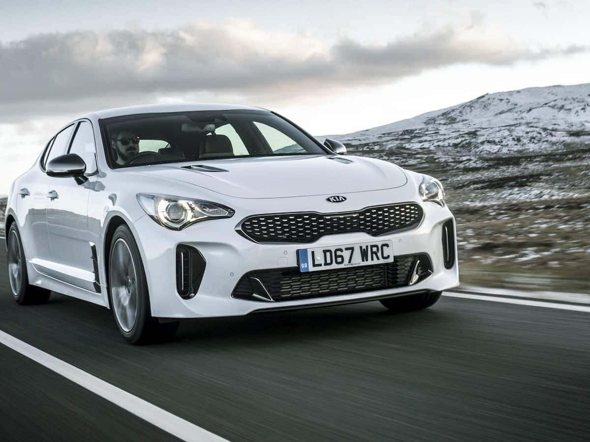Kia Stinger 桌布 壁紙 — 一輛流線型白色Kia Stinger在蜿蜒的道路上疾馳，背景是白雪覆蓋的山脈，天空多雲。