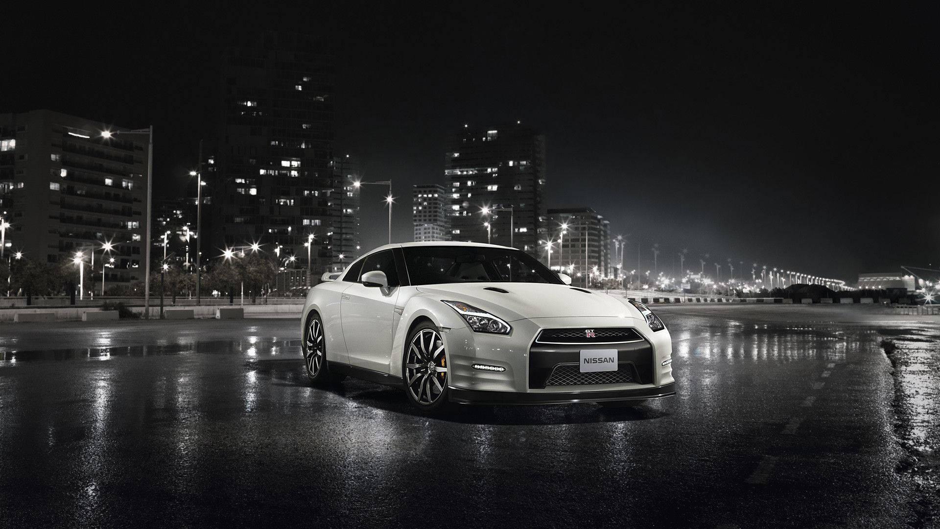 Nissan 桌布 壁紙 — 一輛流線型白色日產GT-R在夜間濕漉漉的瀝青路上，於城市燈光下閃耀，背景是高樓大廈發出的光芒。