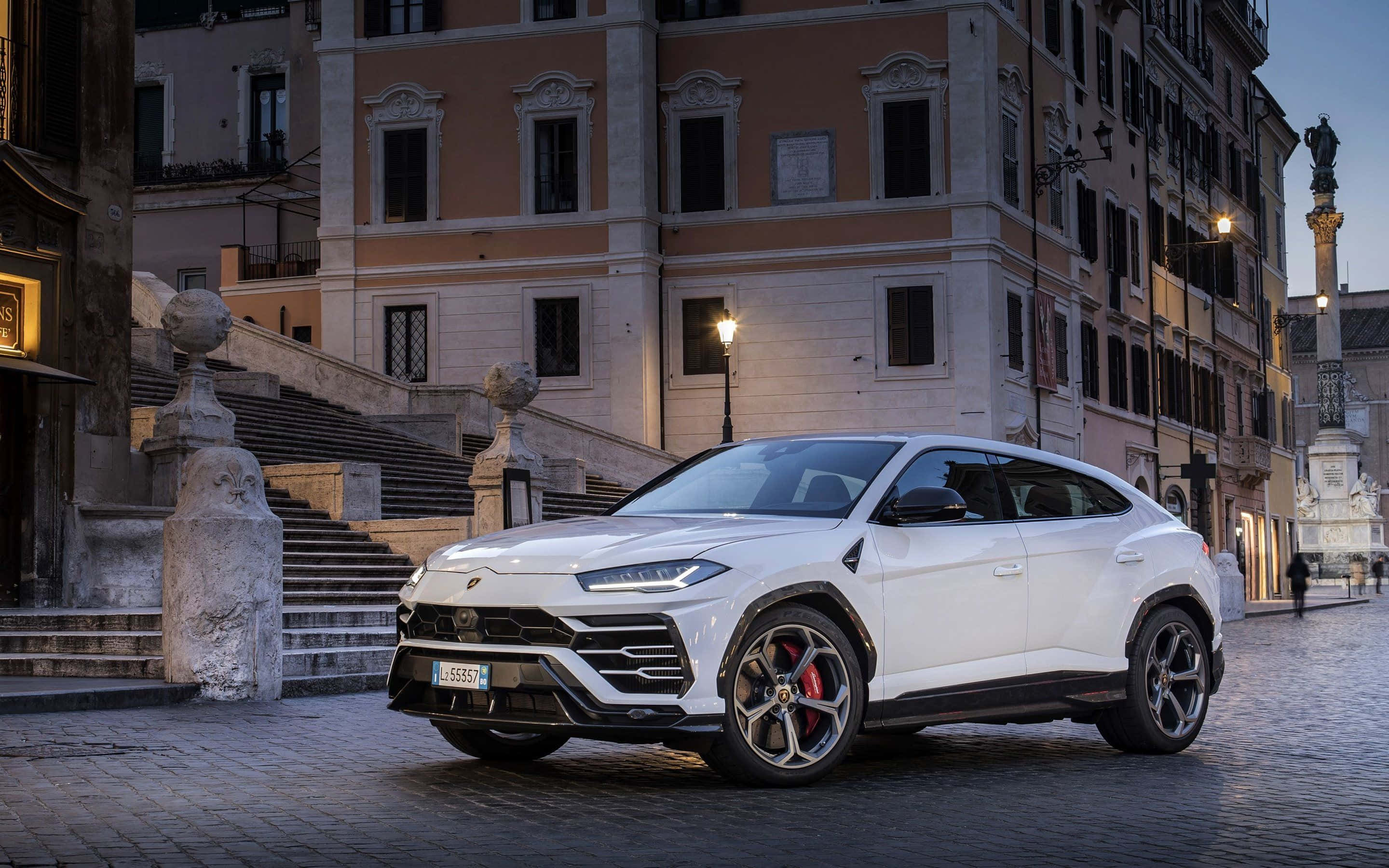 Lamborghini Urus 桌布 壁紙 — 一輛流線型白色藍寶堅尼Urus SUV停泊在黃昏的鵝卵石街道上，背景是歷史悠久的歐洲建築和溫暖的路燈。