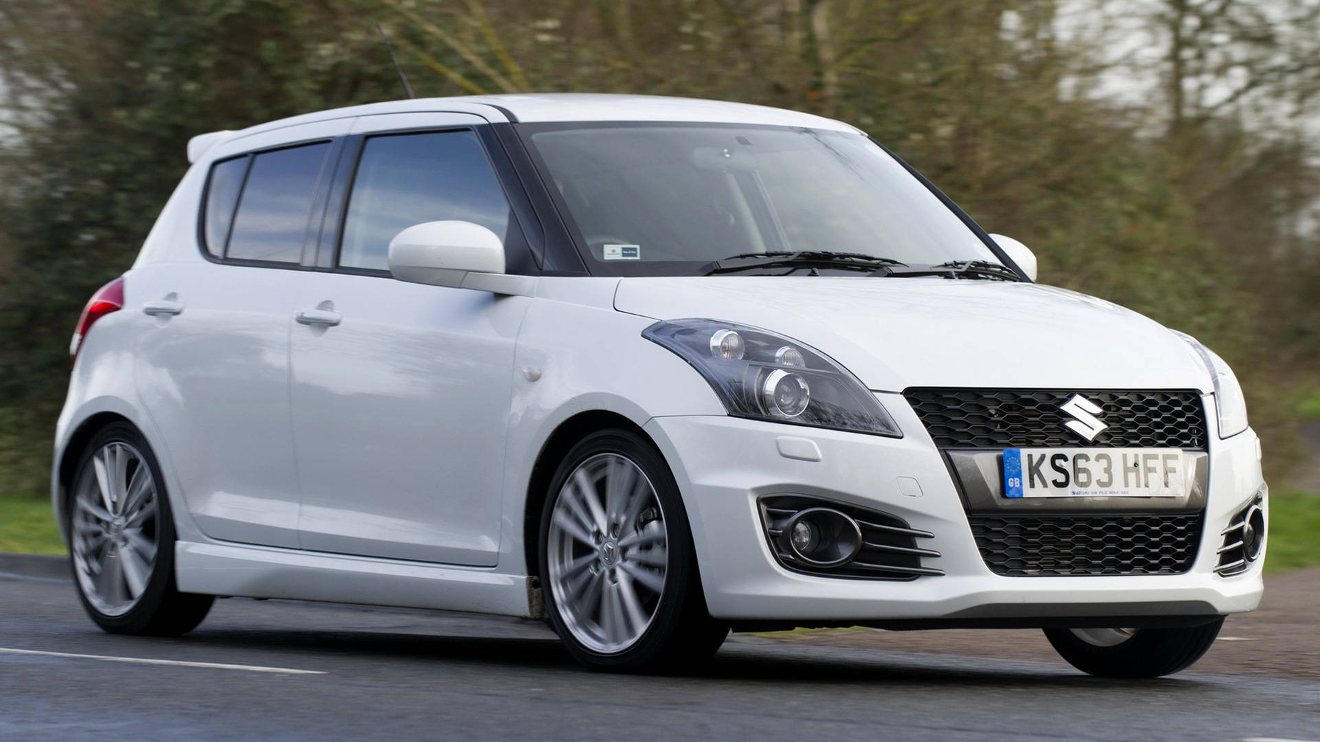 Suzuki 桌布 壁紙 — 一輛流線型白色鈴木 Swift 掀背車在道路上行駛，背景有模糊的綠色樹木。