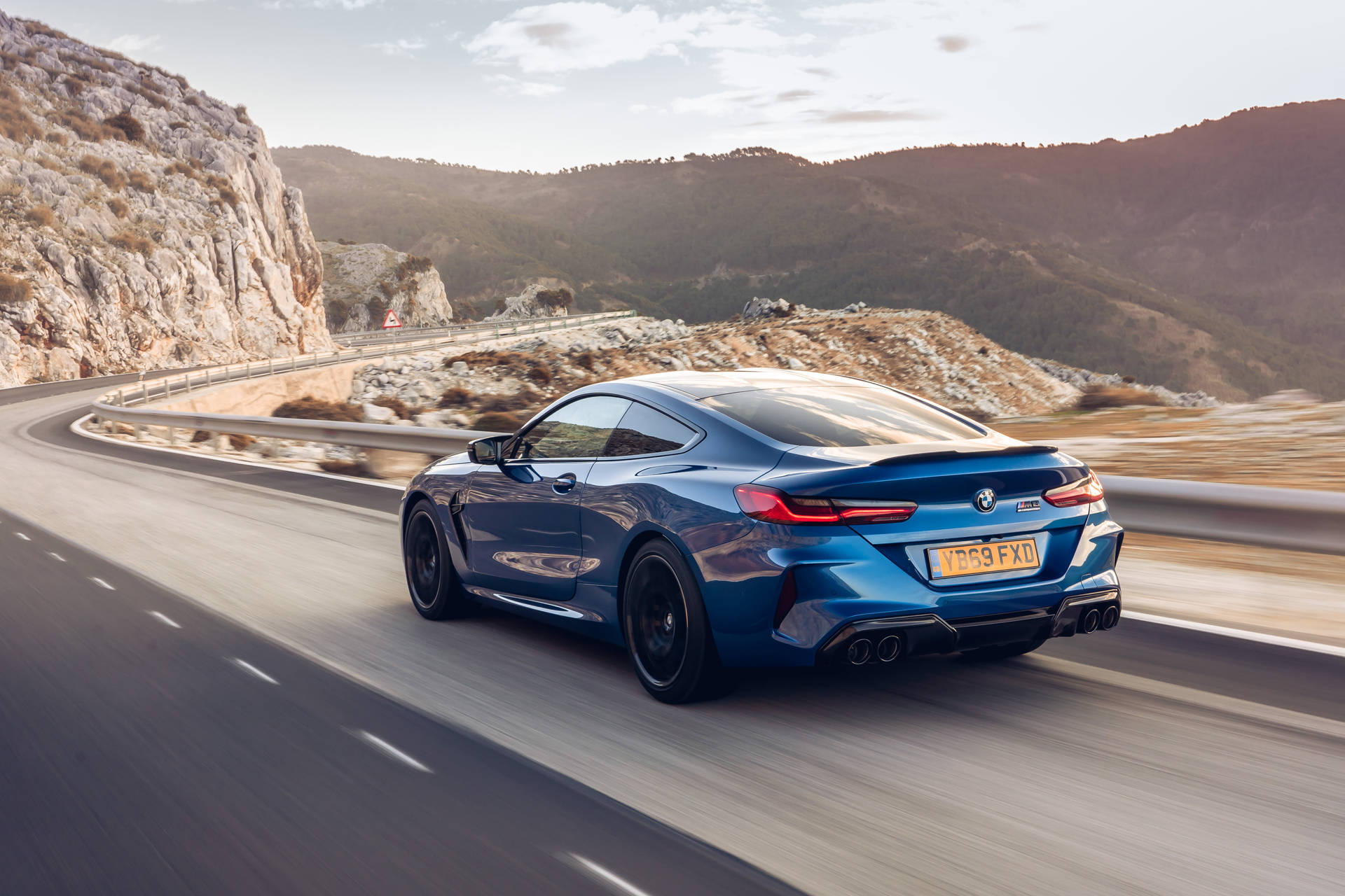 BMW 動圖 桌布 壁紙 — 一輛流線型的藍色BMW M8 Coupe在蜿蜒的山路上疾馳，沿途有岩石峭壁和遠山，天空呈現柔和的金色。