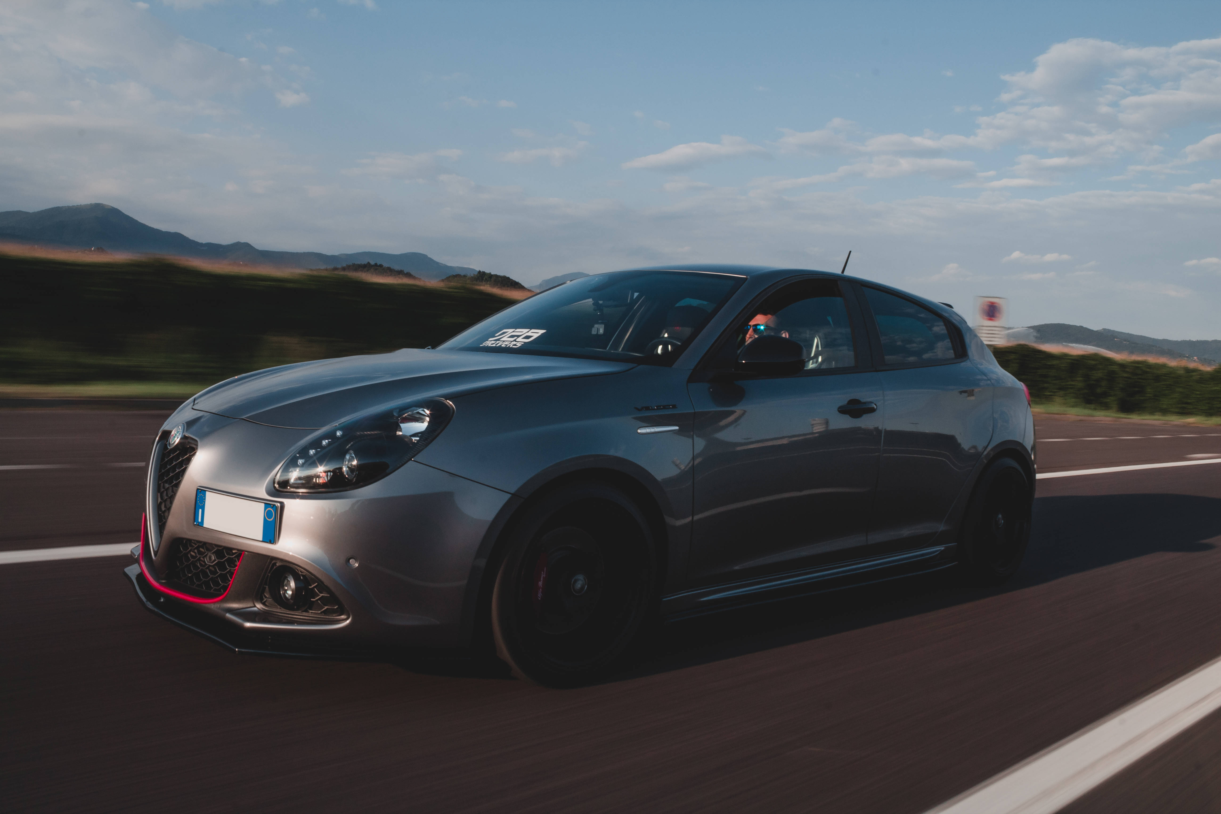 Alfa Romeo 桌布 壁紙 — 一輛流線型的銀色愛快羅密歐Giulietta Veloce在蜿蜒的道路上飛馳，背景是模糊的綠色山丘和部分多雲的天空。