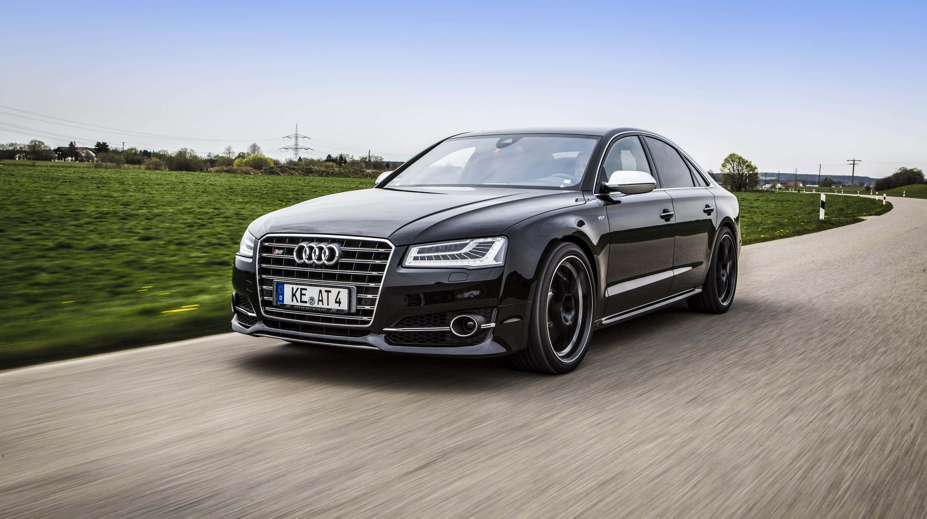 Audi S8 桌布 壁紙 — 一輛流線型的黑色 Audi 房車在蜿蜒的鄉間道路上疾馳，背景是模糊的綠色田野和清澈的藍天。