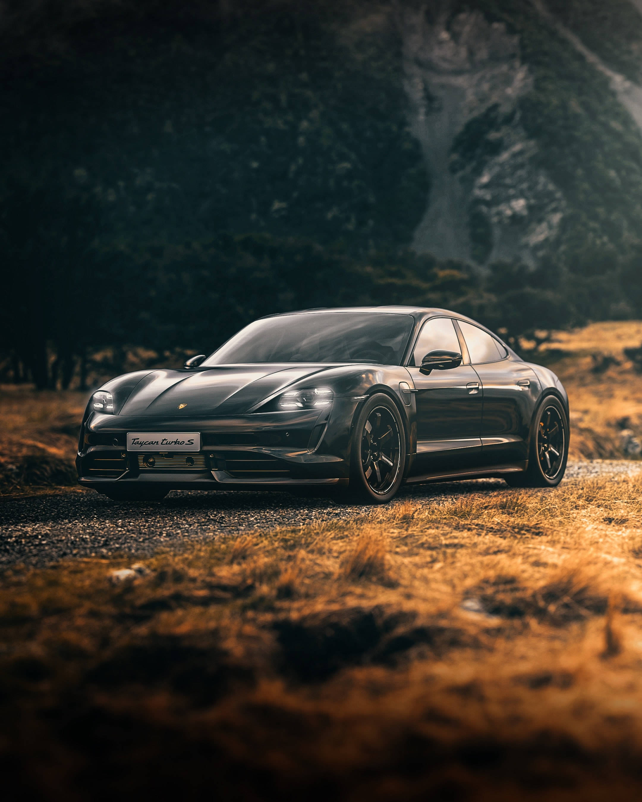 汽車 桌布 壁紙 — 一輛流線型的黑色保時捷 Taycan Turbo S 在金色夕陽下閃耀於碎石路上，背景是森林覆蓋的山脈。