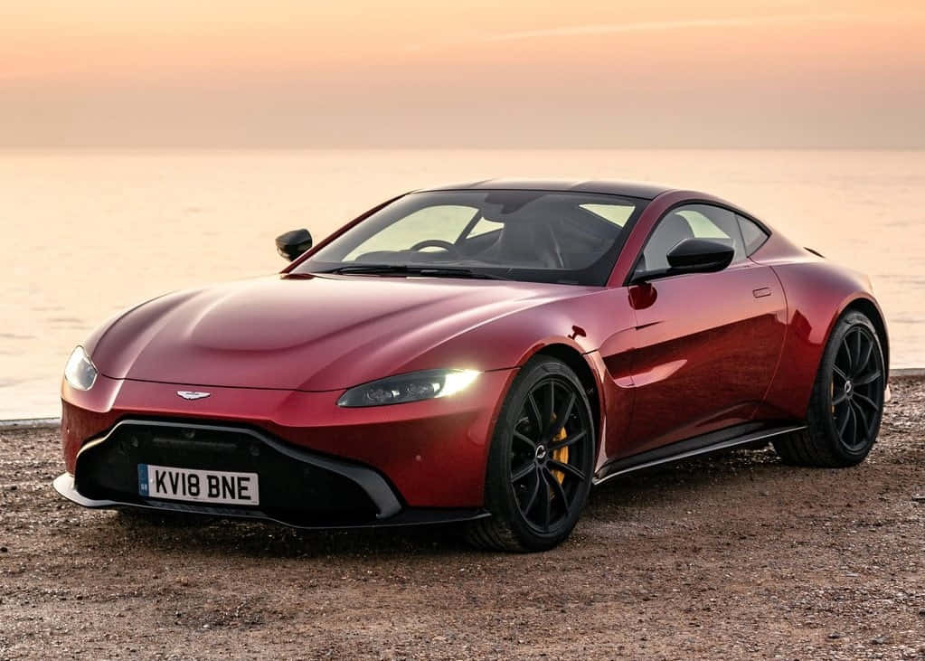 奧斯頓馬丁 桌布 壁紙 — 一輛流線型紅色Aston Martin跑車在日落時分，於卵石海灘上閃耀，背後是平靜的海水。