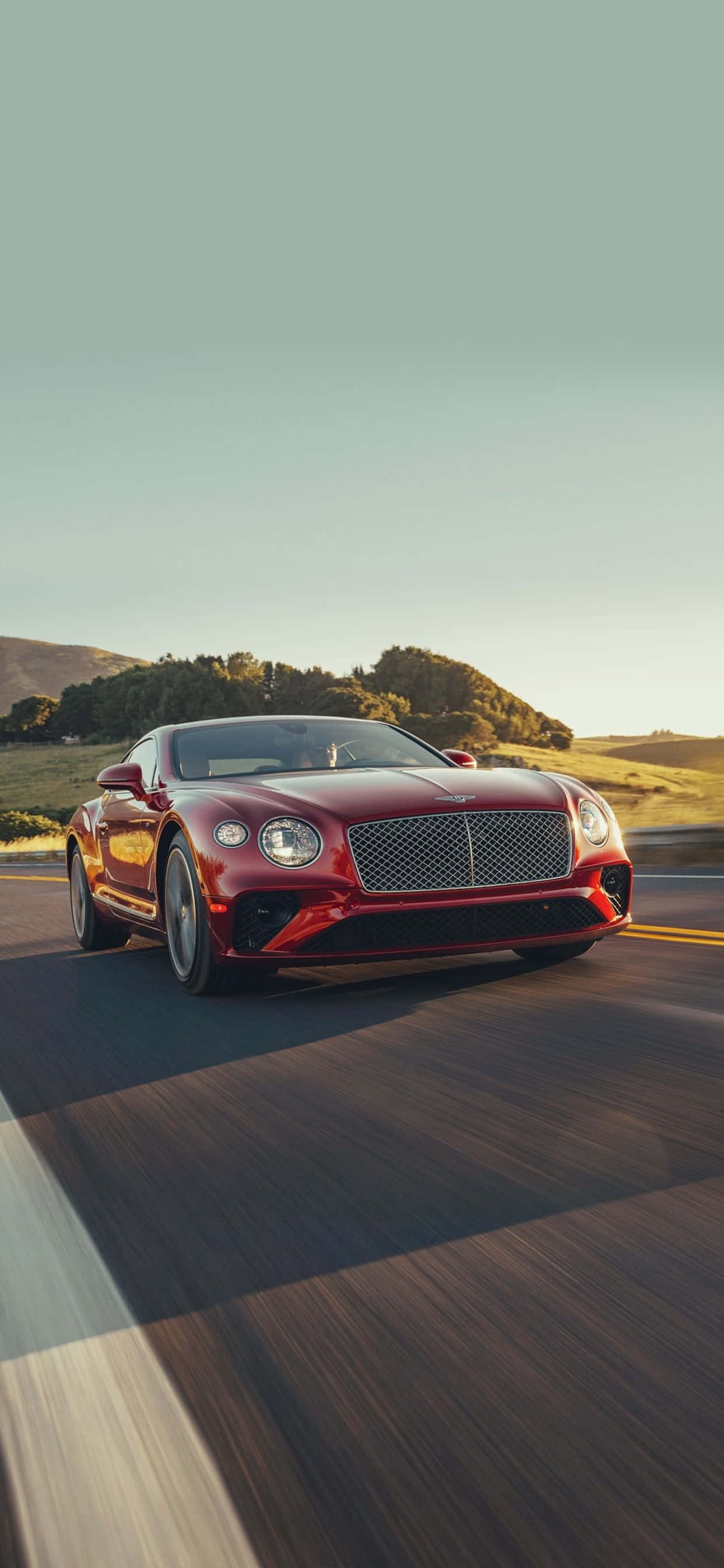 iPhone Xs Max Bentley 桌布 壁紙 — 一輛流線型紅色賓利 Continental GT 在晴朗天空下沿著蜿蜒道路穿過金色山丘飛馳。