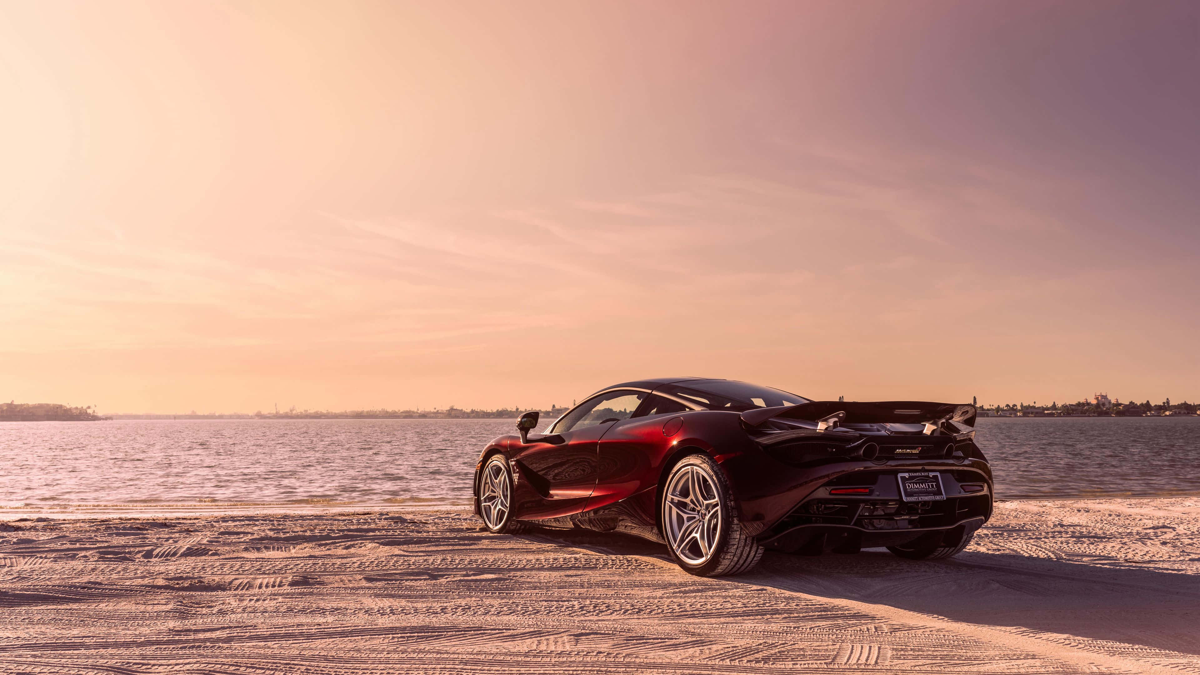 4K Mclaren 720s 桌布 壁紙 — 一輛流線型紅色跑車停泊在雪灘上，夕陽西下，平靜的海洋和遠處的城市天際線。