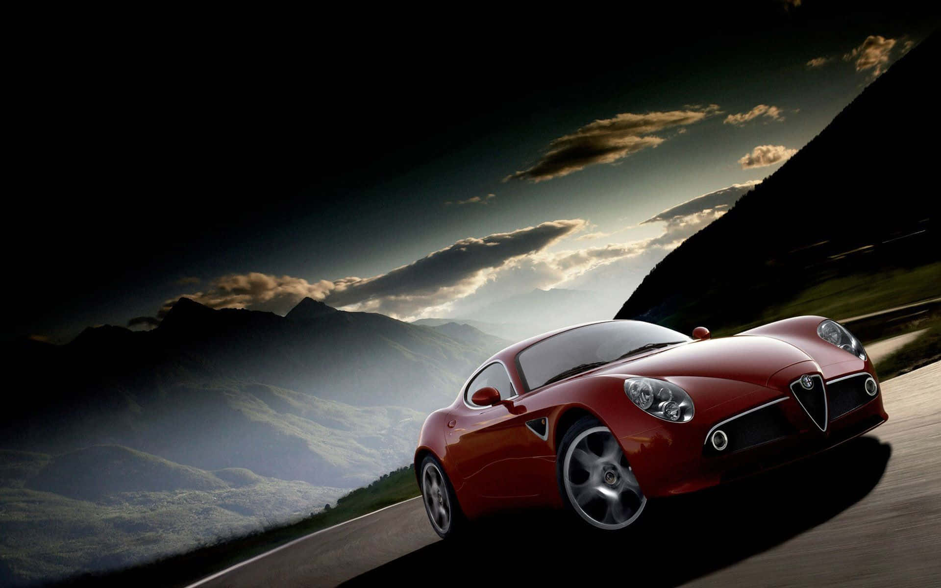 Alfa Romeo 8c Competizione 桌布 壁紙 — 一輛流線型紅色跑車在日落時分沿著蜿蜒的山路疾馳，背景是戲劇性的雲彩和霧氣繚繞的山峰。