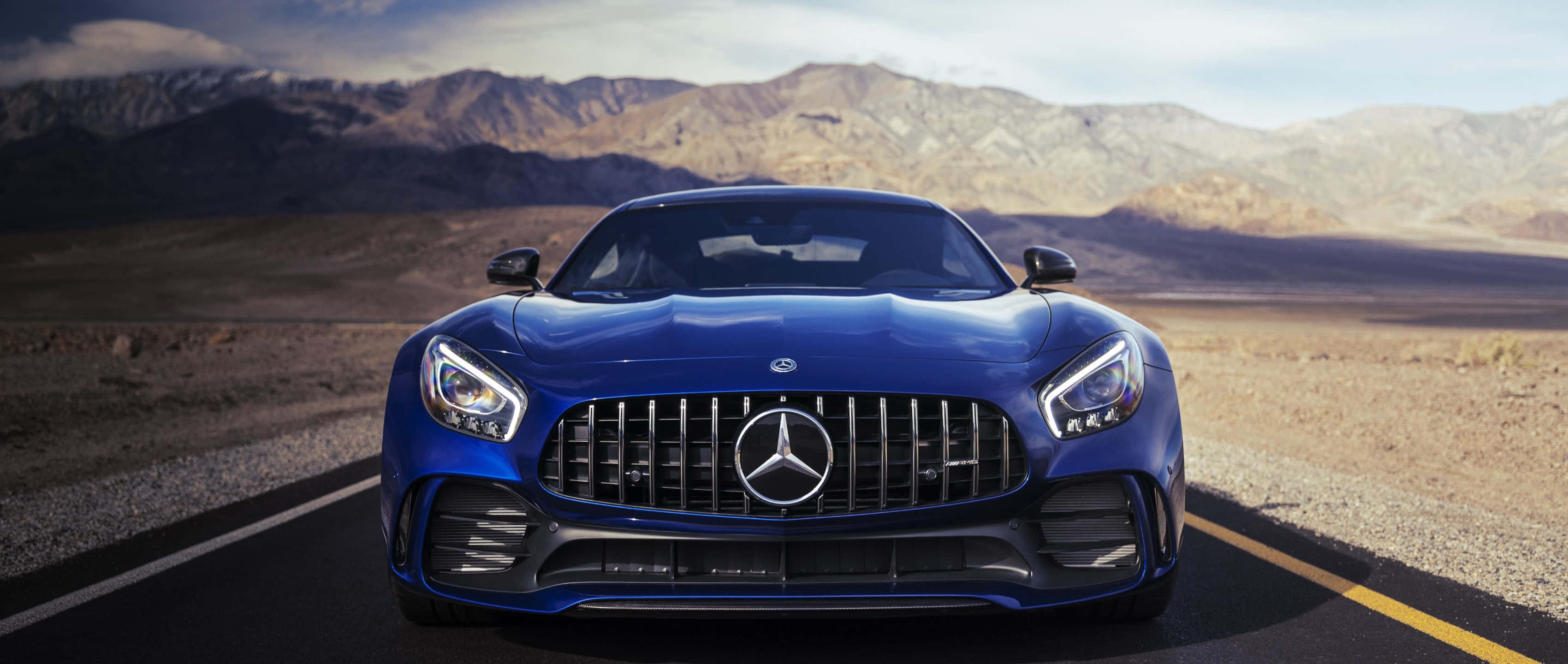 Mercedes 桌布 壁紙 — 一輛流線型藍色 Mercedes-AMG GT 跑車在沙漠公路上向前行駛，背景是崎嶇的山脈，天空明亮。