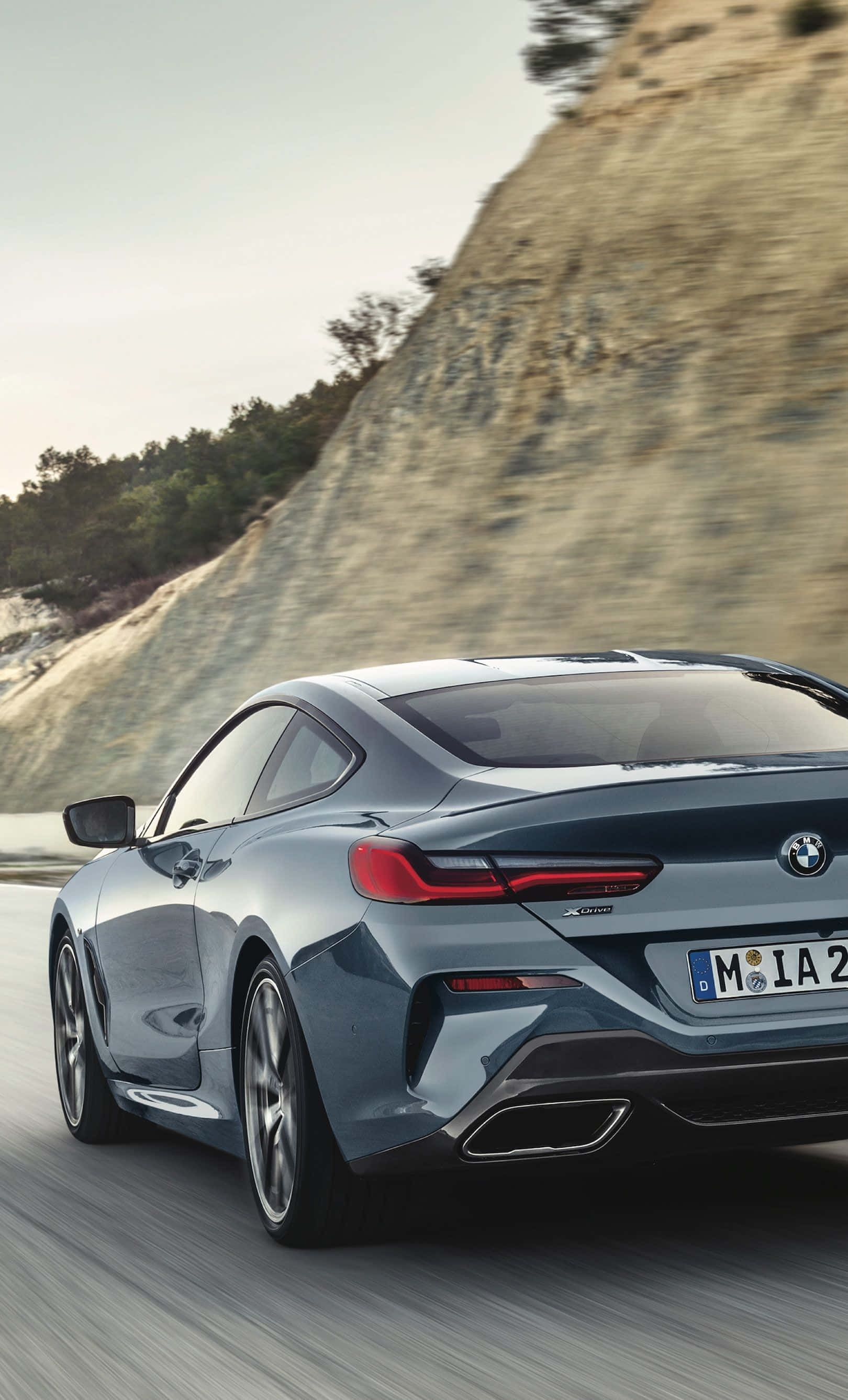 Bmw iPhone 桌布 壁紙 — 一輛流線型藍色BMW 8系列轎跑車在蜿蜒的山路上奔馳，帶有模糊的動感和岩石地形。