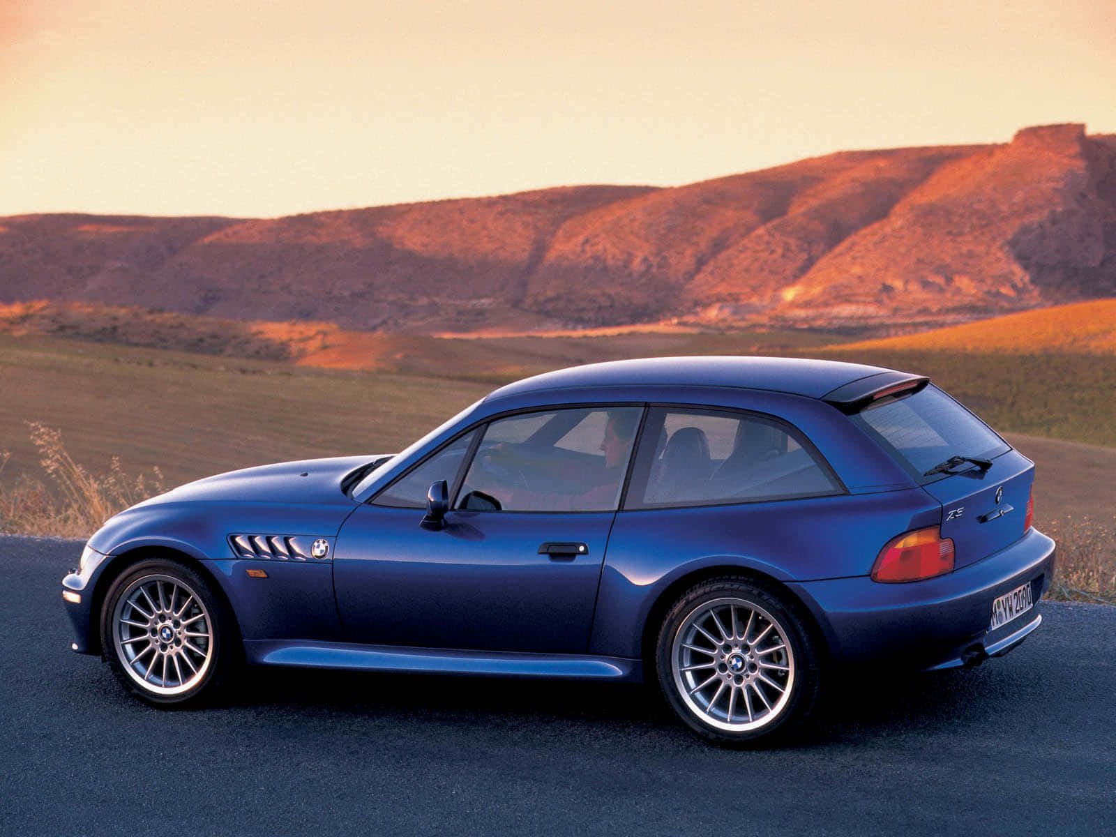 Bmw Z3 桌布 壁紙 — 一輛流線型藍色BMW Z3敞篷跑車停泊在沙漠公路上，背景是夕陽下的連綿山丘。