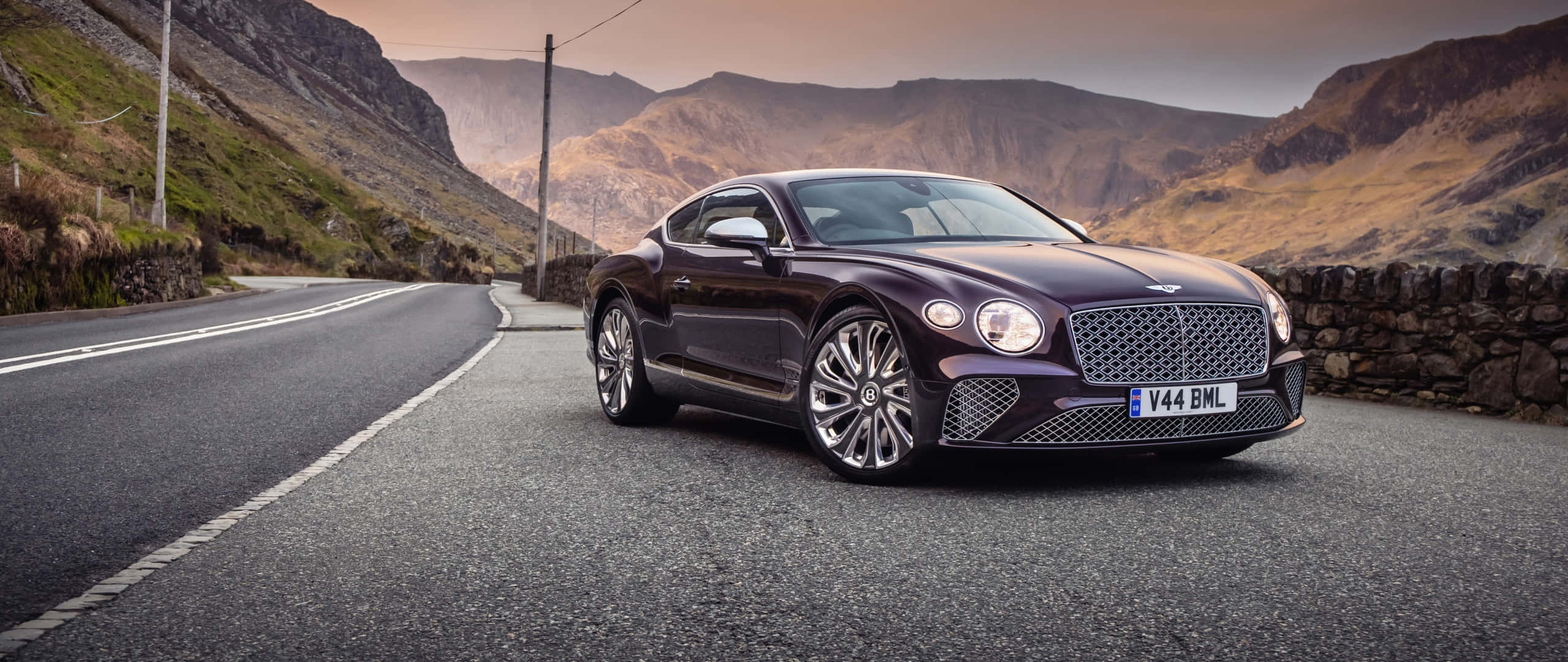 Bentley Mulsanne 桌布 壁紙 — 一輛流線型酒紅色賓利 Continental GT 停泊在蜿蜒的山路上，背景是崎嶇的山丘和石牆，天空呈現柔和的金色。