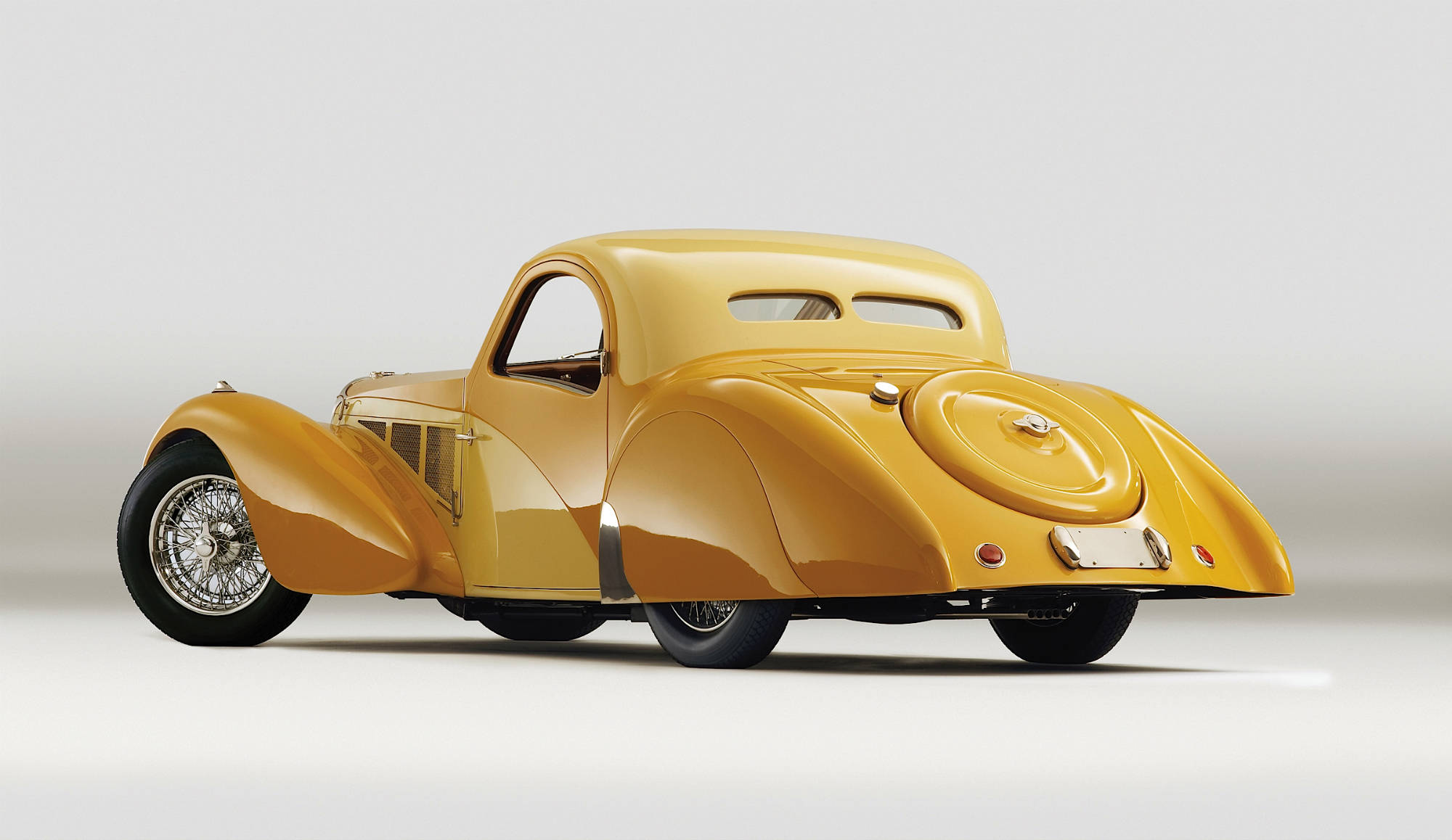 Cool Bugatti 桌布 壁紙 — 一輛流線型金色復古車，帶有流暢曲線和鋼絲輻條輪，置於簡約工作室燈光下。