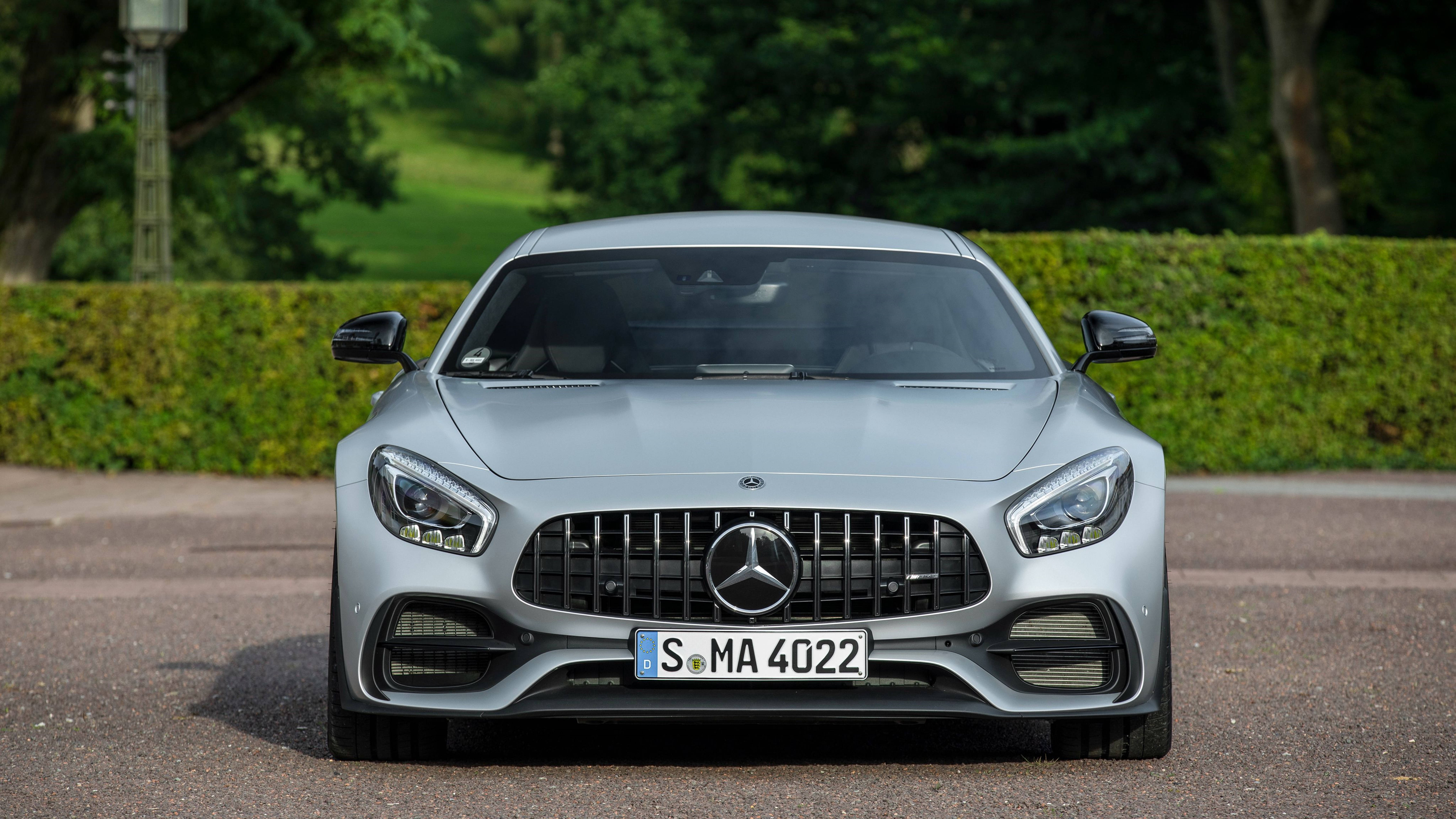 Mercedes 桌布 壁紙 — 一輛流線型銀色 Mercedes-AMG GT 跑車在鋪砌道路上向前行駛，背景有茂密的綠色籬笆和樹木。