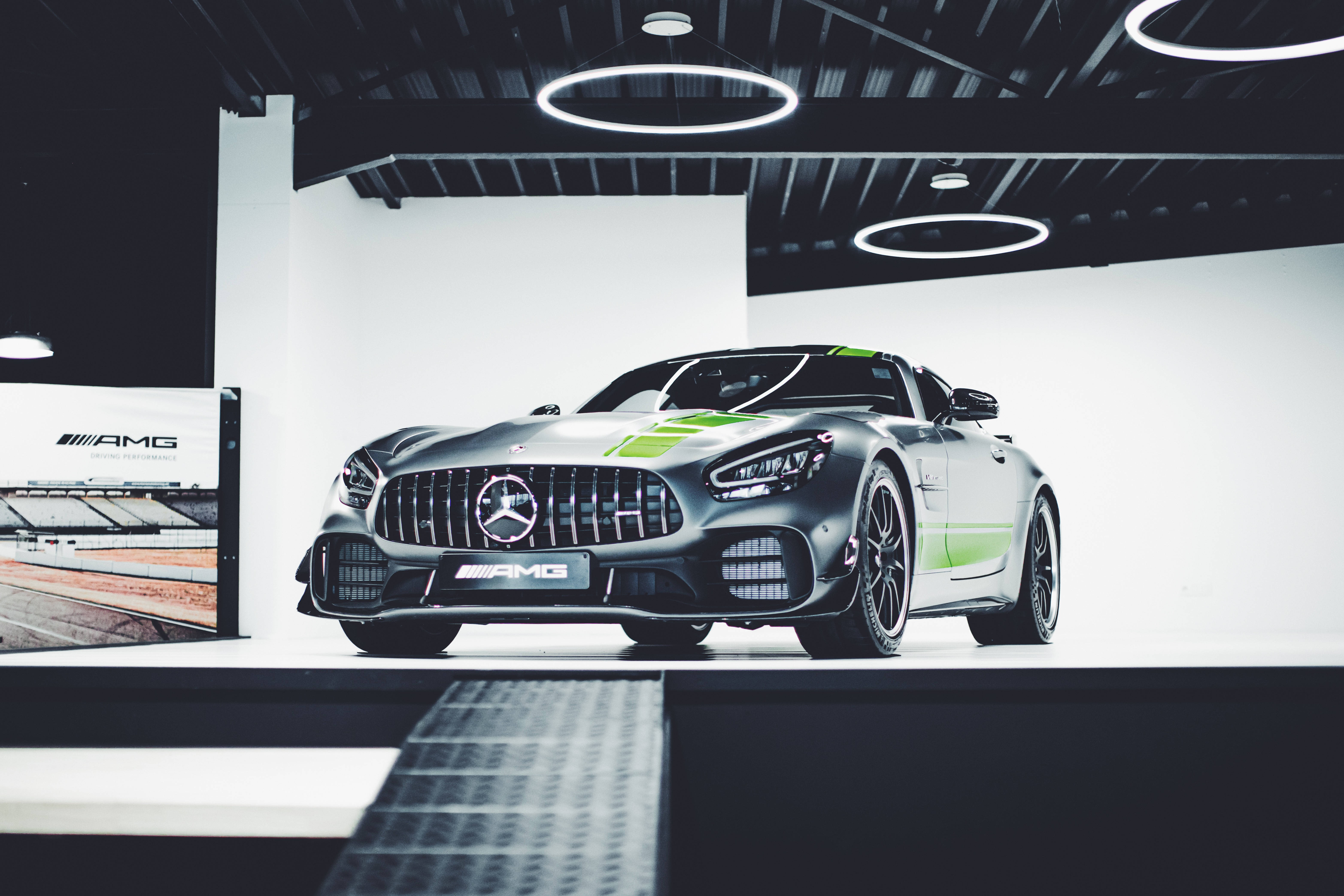 Mercedes Amg 桌布 壁紙 — 一輛流線型銀色AMG GT賽車帶有綠色點綴，在圓形燈光下於極簡工作室環境中展示。