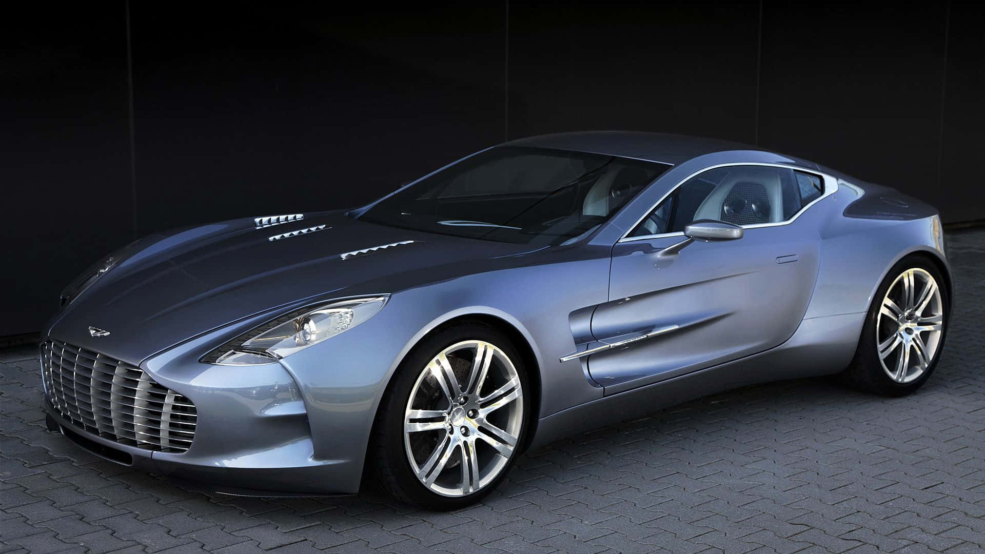 Aston Martin One-77 桌布 壁紙 — 一輛流線型銀色Aston Martin One-77跑車在攝影棚燈光下，於深色鋪砌路面上閃閃發光。