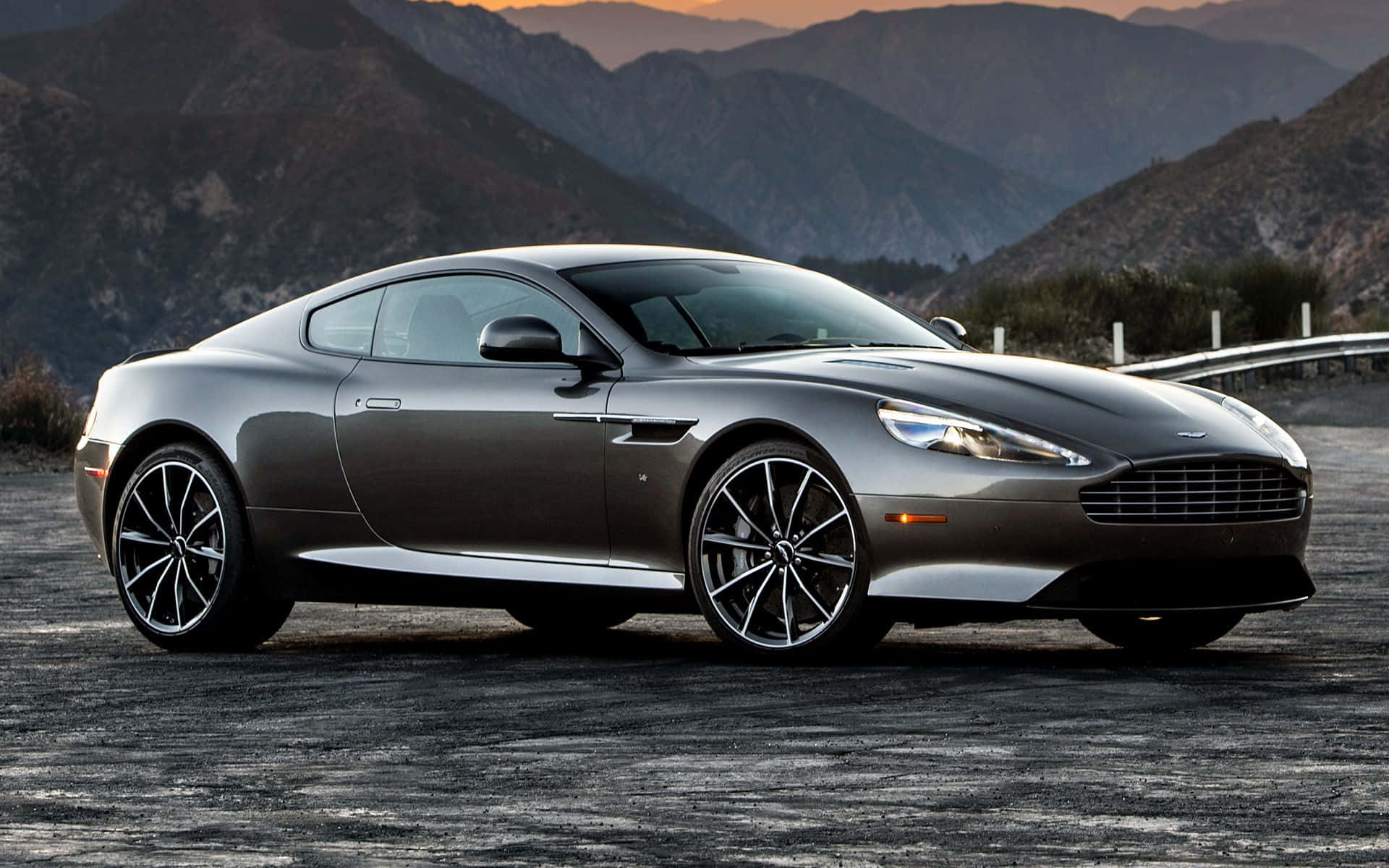 Aston Martin Db9 桌布 壁紙 — 一輛流線型銀色Aston Martin跑車停泊在日落時分的山路上，背景是崎嶇的山峰。