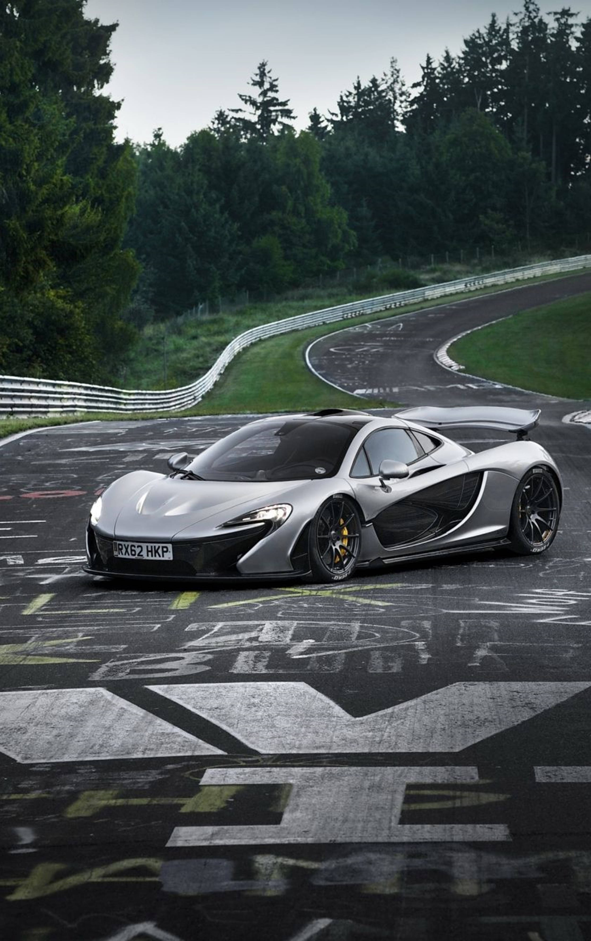 Luxury 桌布 壁紙 — 一輛流線型銀色McLaren P1跑車在多雲的天空下，沿著蜿蜒的瀝青賽道行駛，賽道兩旁是茂密的綠色森林。