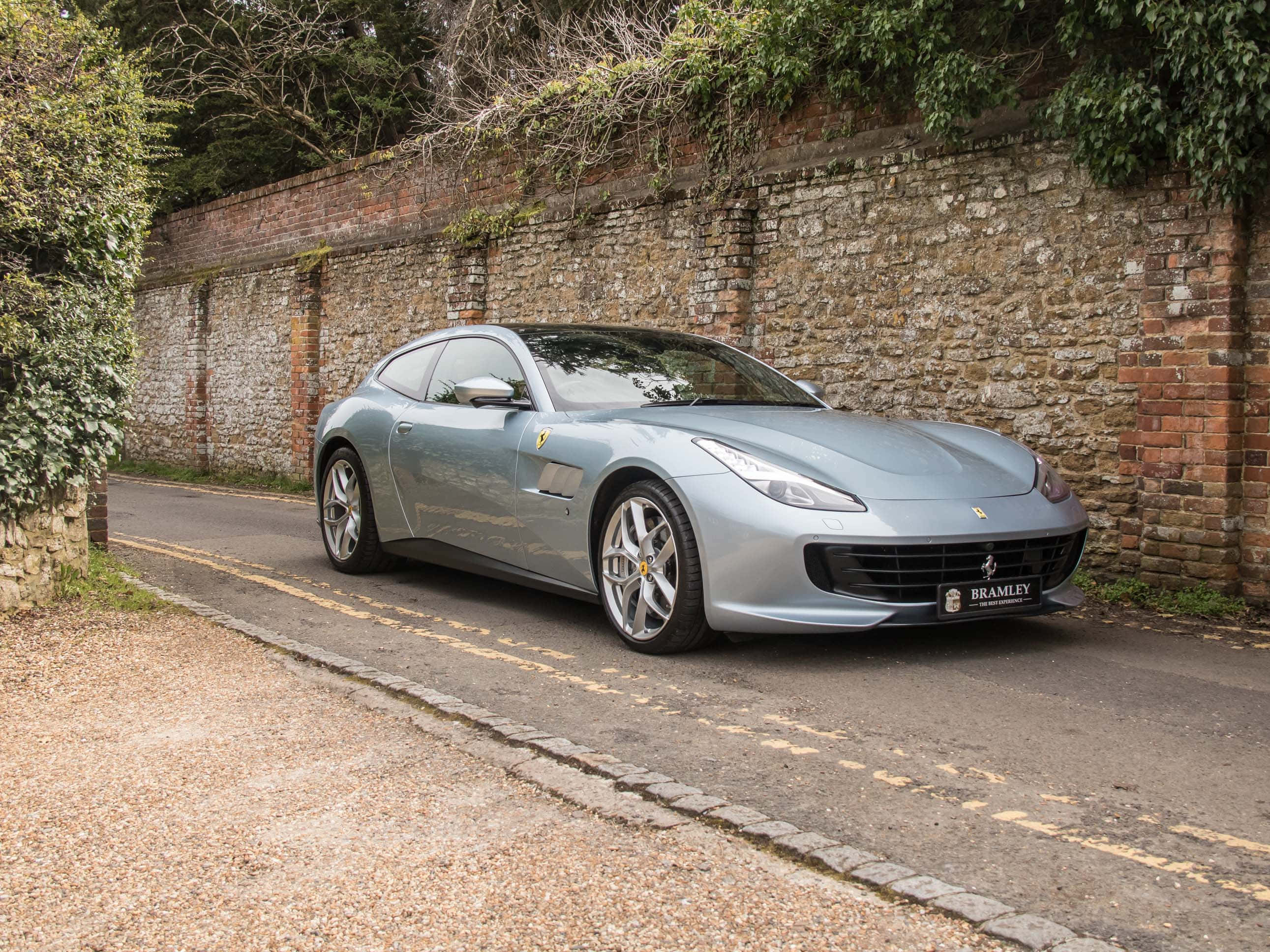 Ferrari Gtc4lusso 桌布 壁紙 — 一輛流線型銀色法拉利 GTC4Lusso 停泊在寧靜的道路上，旁邊是長滿青苔的石牆，常春藤攀爬而上。