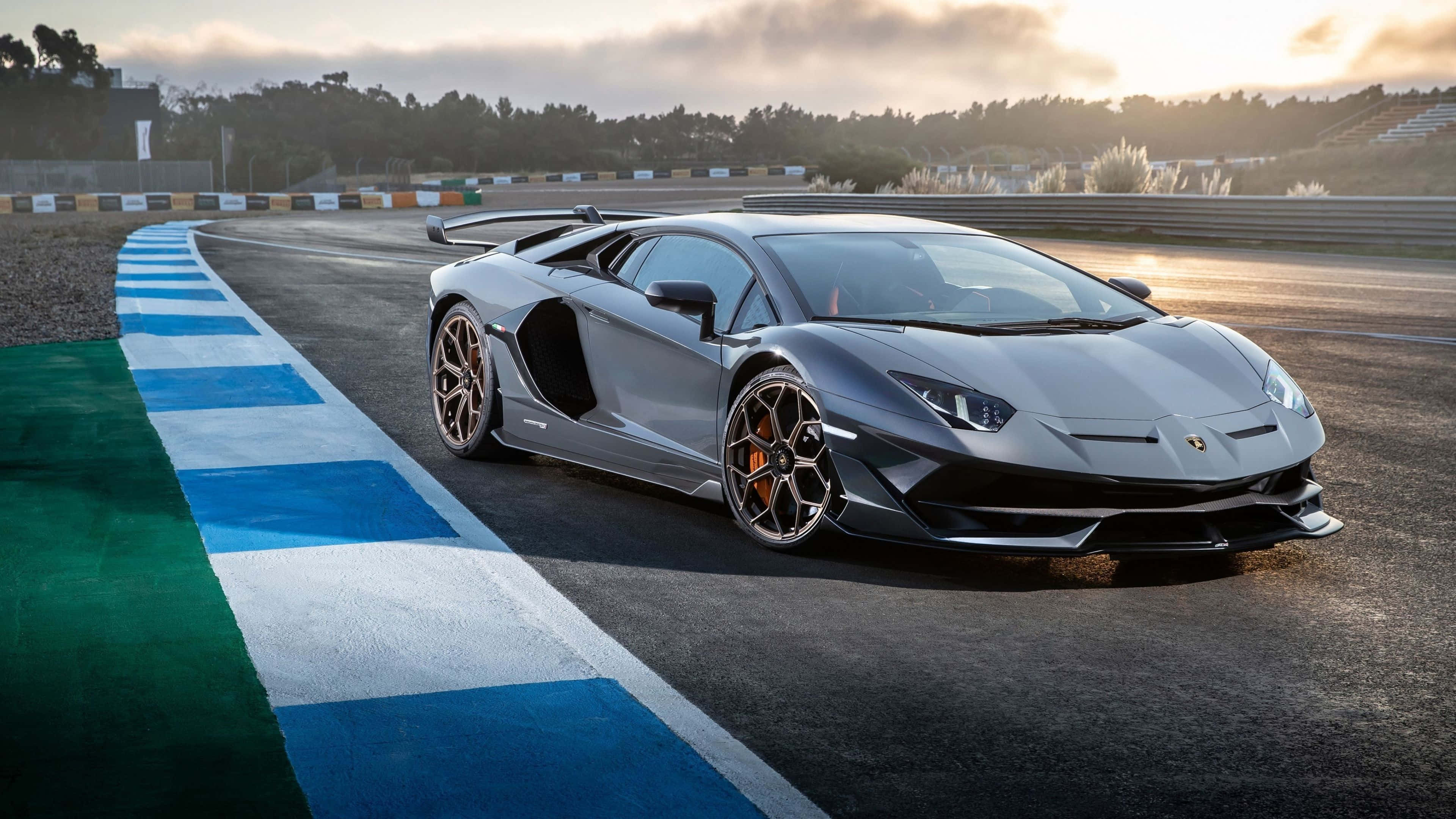 4K 藍寶堅尼 桌布 壁紙 — 一輛流線型銀色藍寶堅尼 Aventador SVJ 在陽光明媚的賽道上奔馳，賽道有藍白路緣石，遠處有看台，天空多雲。