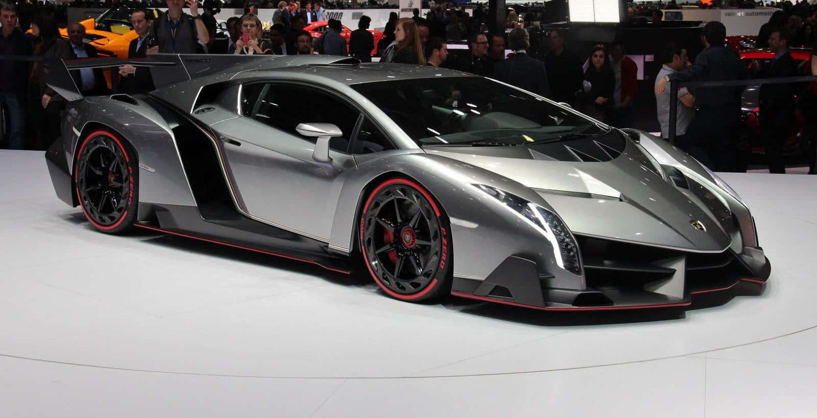 Lamborghini Veneno 桌布 壁紙 — 一輛流線型銀色藍寶堅尼 Veneno 搭配紅色輪圈，在車展上展出，周圍人群簇擁。