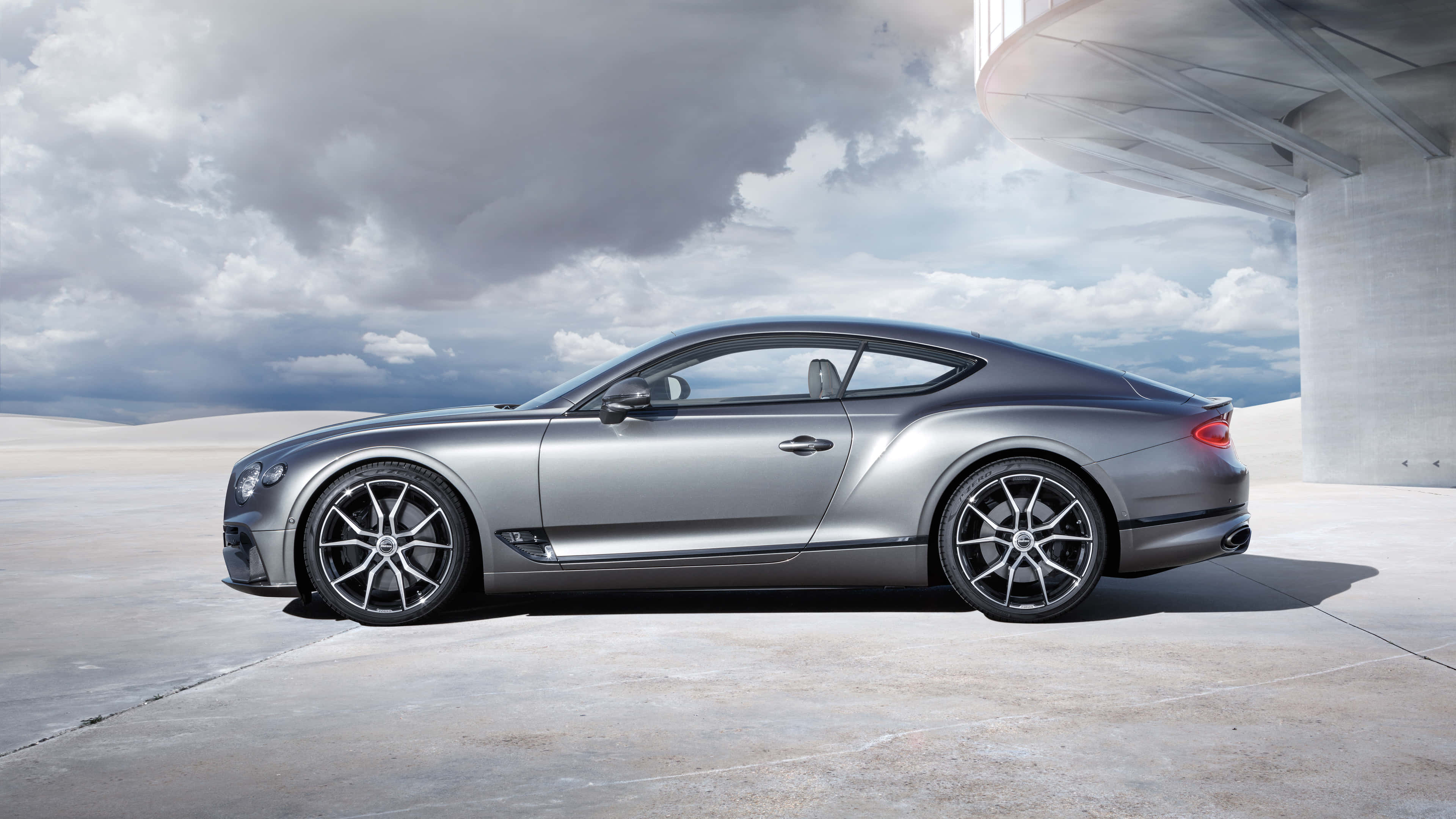 Bentley Continental Gt 桌布 壁紙 — 一輛流線型銀色賓利 Continental GT 雙門跑車停放在現代混凝土結構下，背景為多雲的天空。