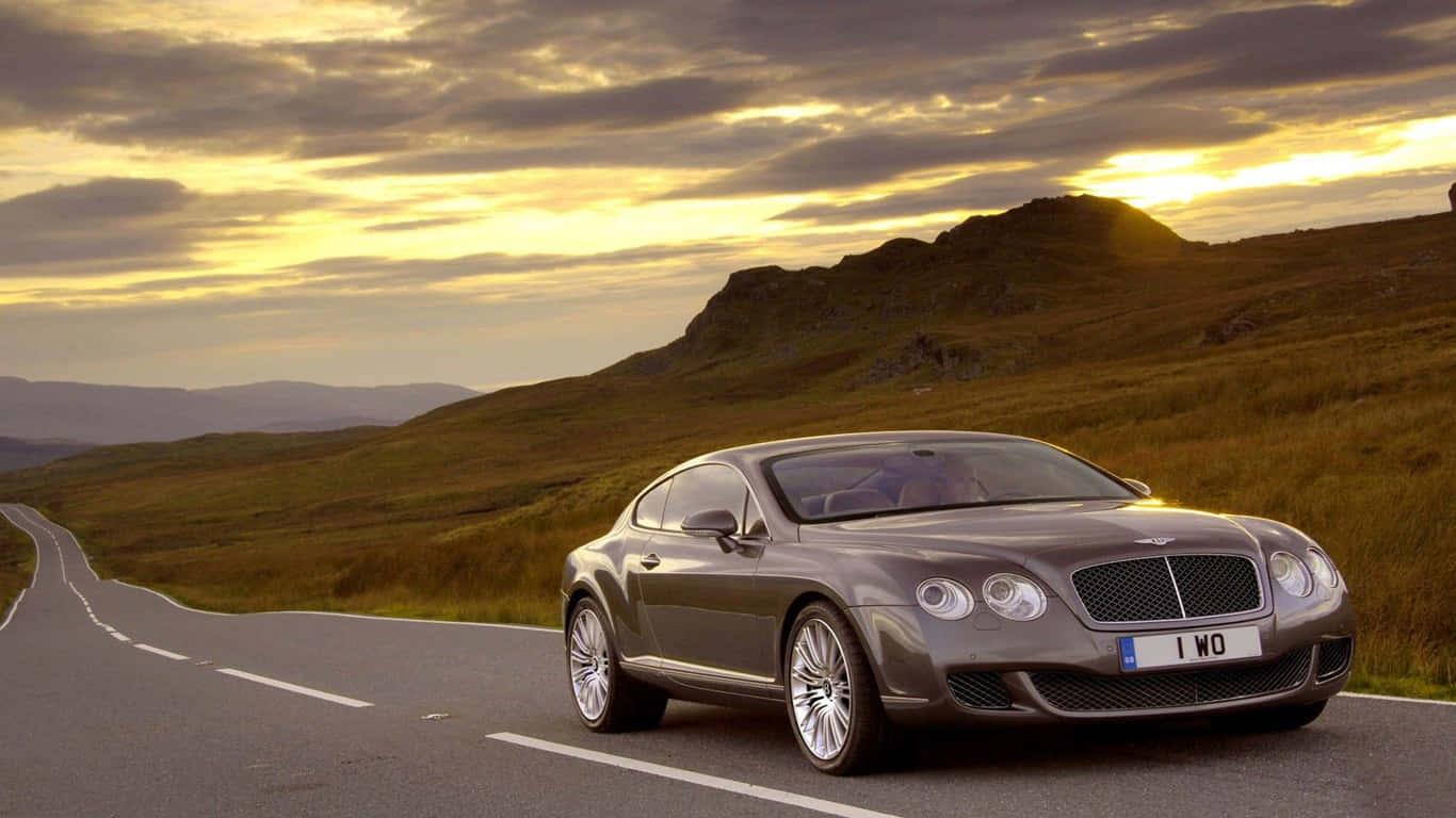 1366x768 Bentley 桌布 壁紙 — 一輛流線型銀色賓利轎跑車沿著蜿蜒的山路行駛，在金色夕陽和戲劇性的雲彩下。
