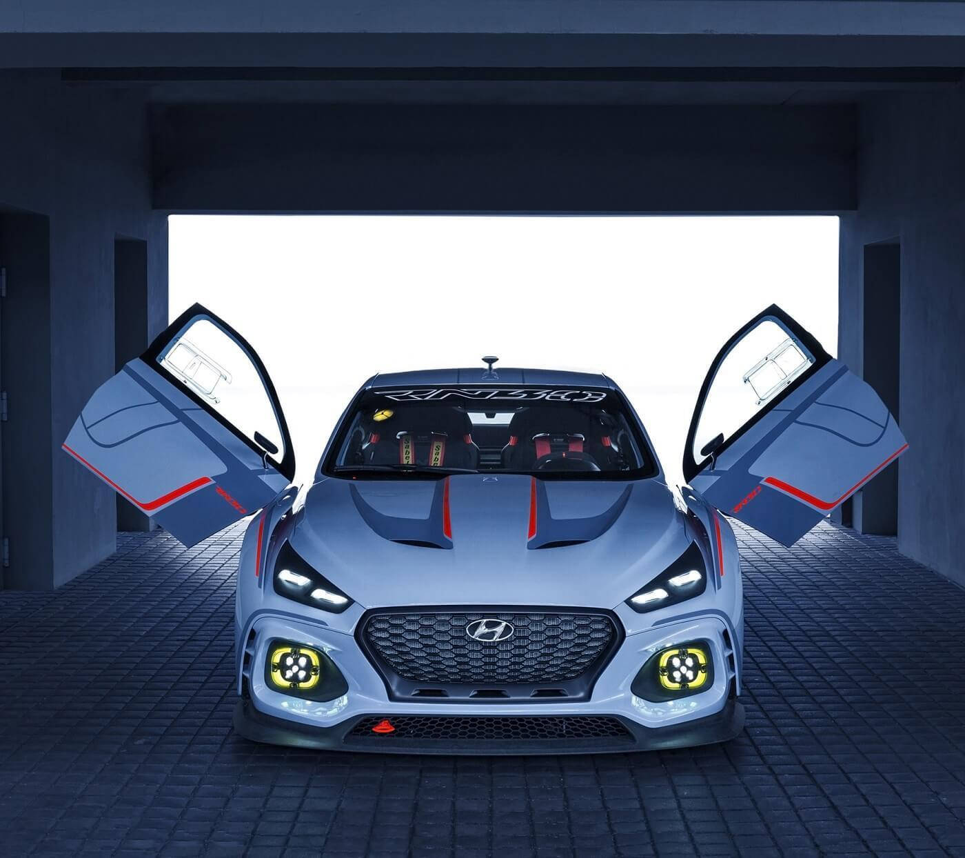 Hyundai 桌布 壁紙 — 一輛流線型銀色賽車打開鷗翼門，停在昏暗的混凝土車庫中，黃色車頭燈照亮鵝卵石地板。