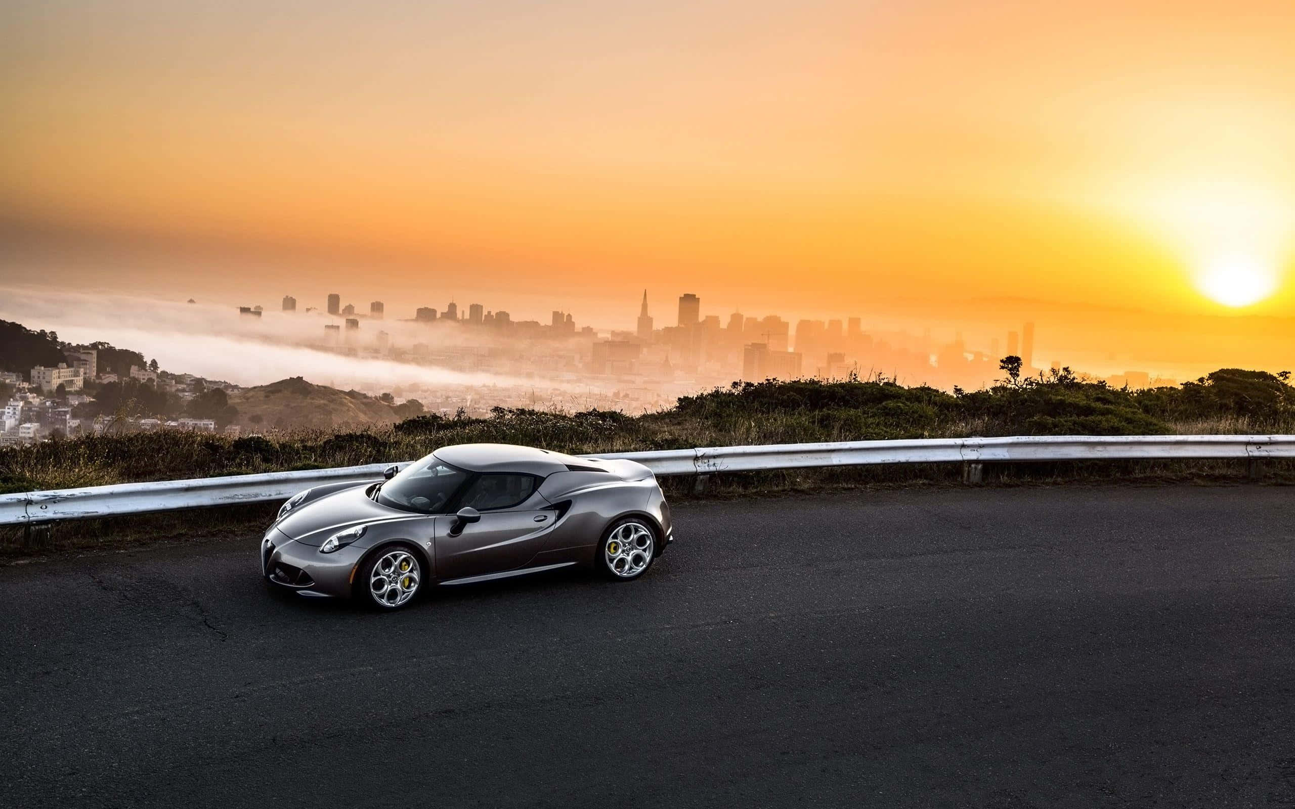 Alfa Romeo 4c 桌布 壁紙 — 一輛流線型銀色跑車停泊在蜿蜒道路上，俯瞰著金色日落時分霧氣朦朧的城市天際線。