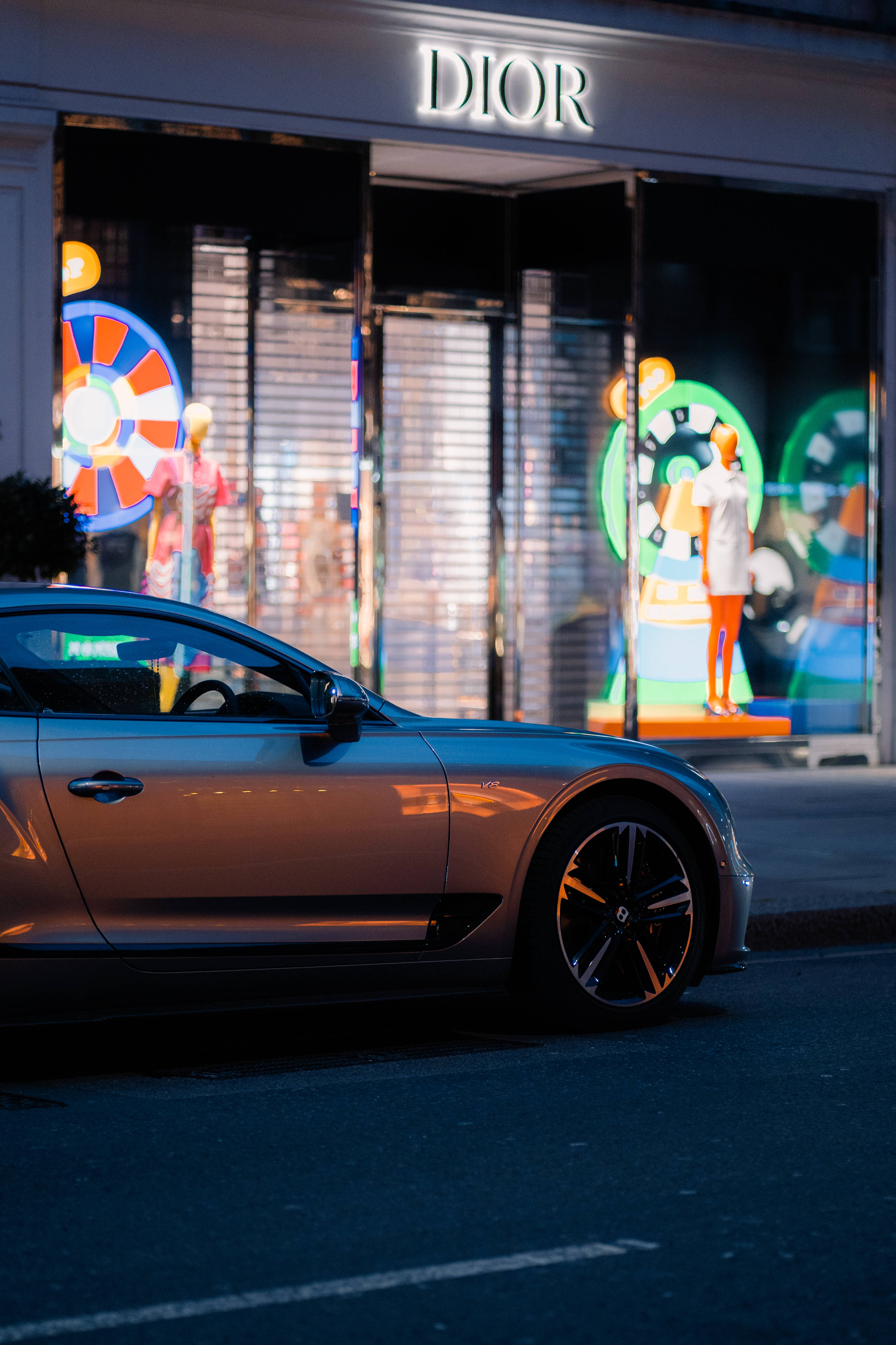 bentley HD 桌布 壁紙 — 一輛流線型銀色跑車在夜間停泊在燈火通明的Dior商店前，櫥窗內有色彩繽紛的人體模型。