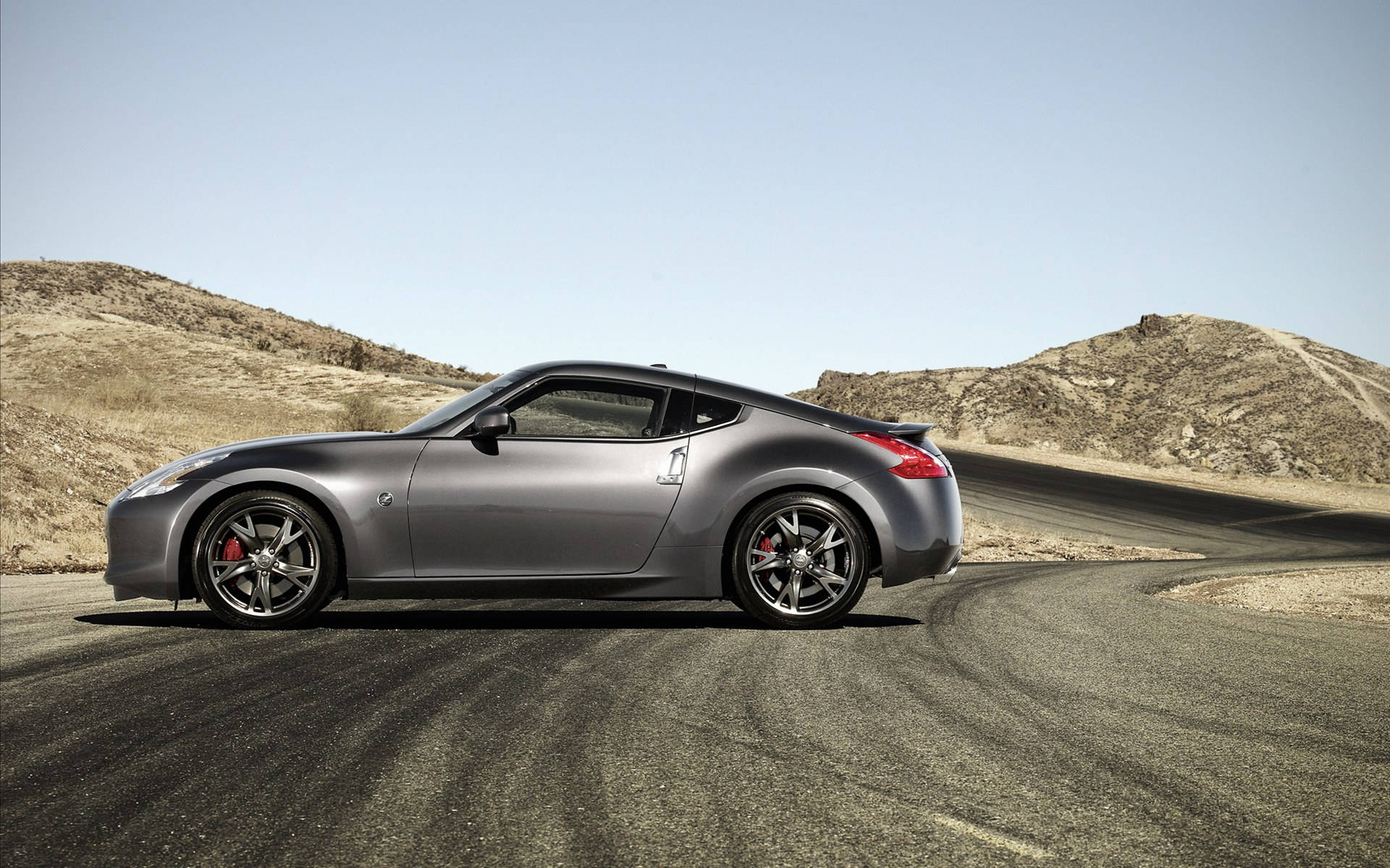Nissan 370z 桌布 壁紙 — 一輛流線型銀色跑車配紅色制動卡鉗，沿著沙漠公路蜿蜒而行，兩旁是乾燥的山丘，頭頂是清澈的藍天。