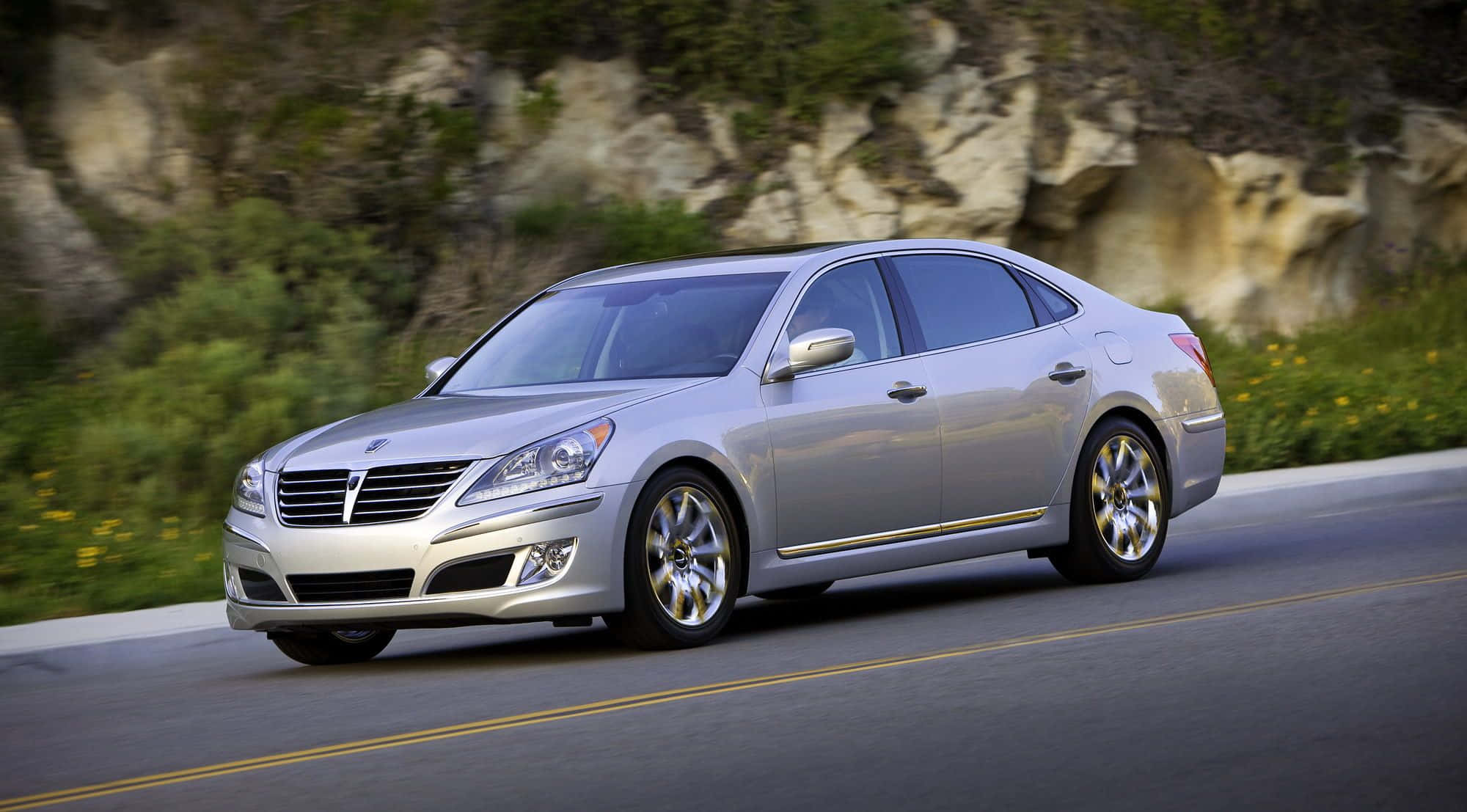 Hyundai Equus 桌布 壁紙 — 一輛流線型銀色轎車沿著蜿蜒的道路疾馳，背景是模糊的綠意和岩石峭壁。