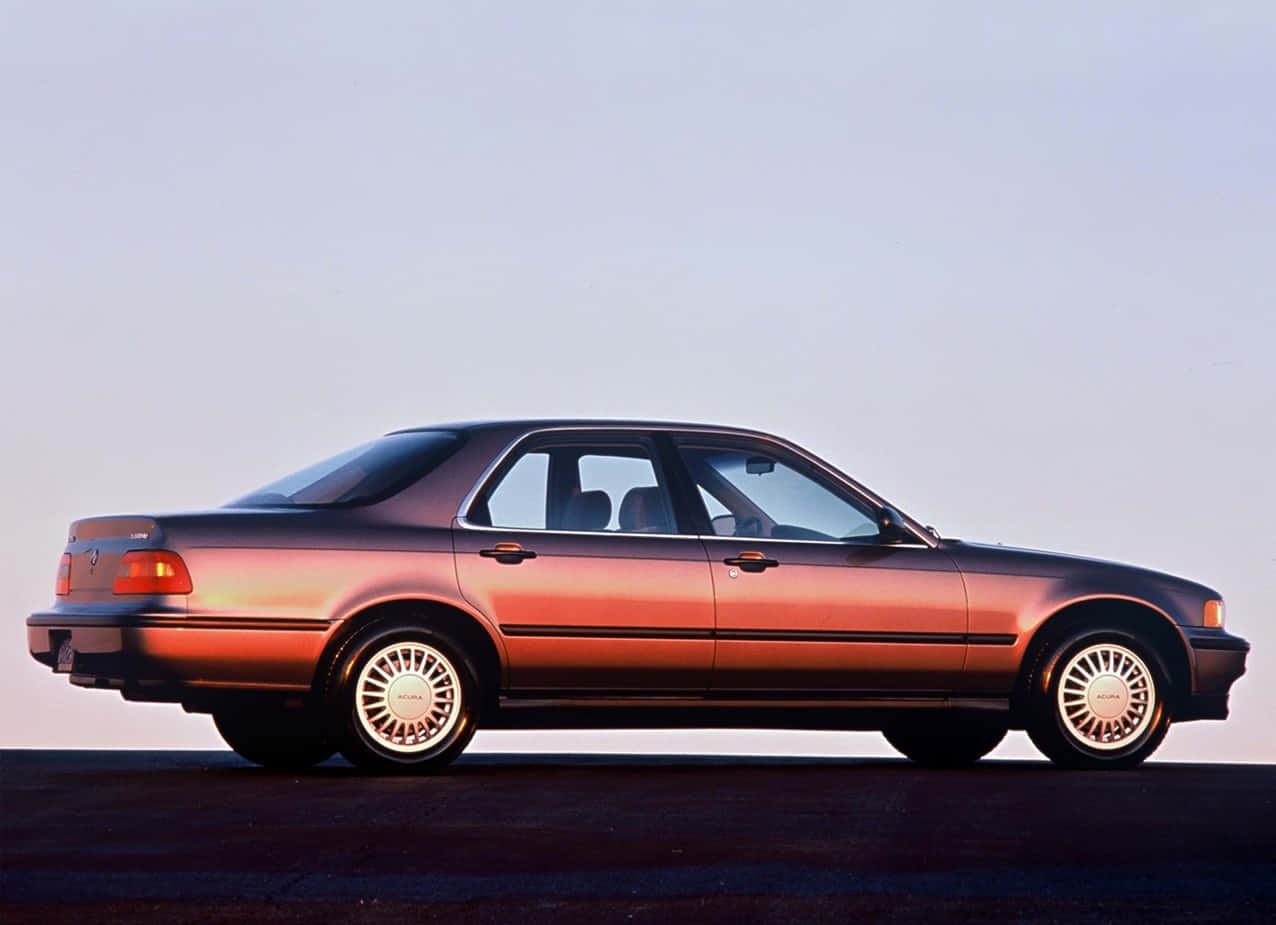 Acura Legend 桌布 壁紙 — 一輛流線型青銅色轎車在柔和暮色天空下以側面輪廓滑行，伴隨溫暖的環境照明。