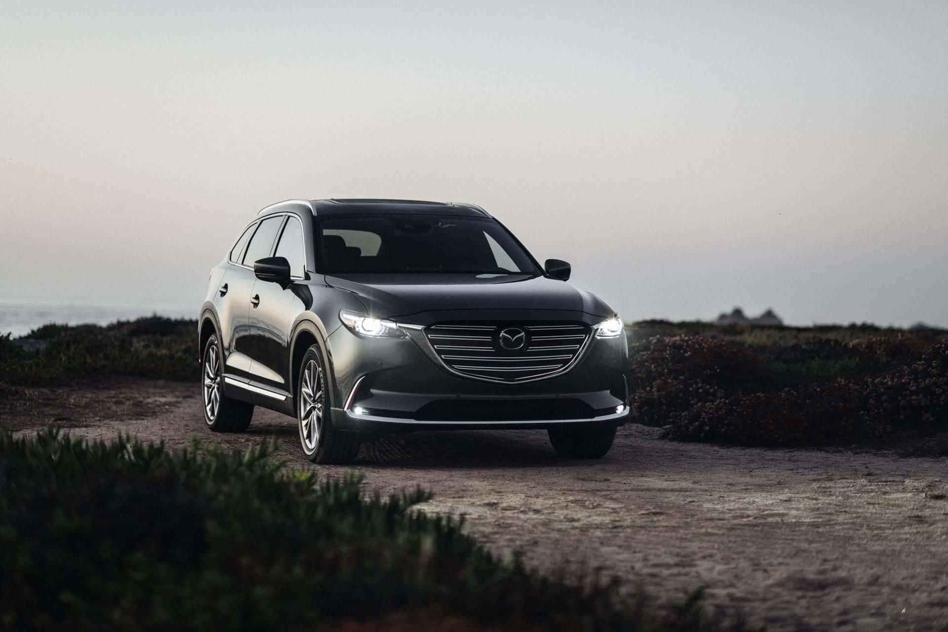 Mazda Cx-9 桌布 壁紙 — 一輛流線型黑色Mazda CX-9 SUV停泊在海岸泥路上，車頭燈亮起，映襯著柔和的暮色天空。
