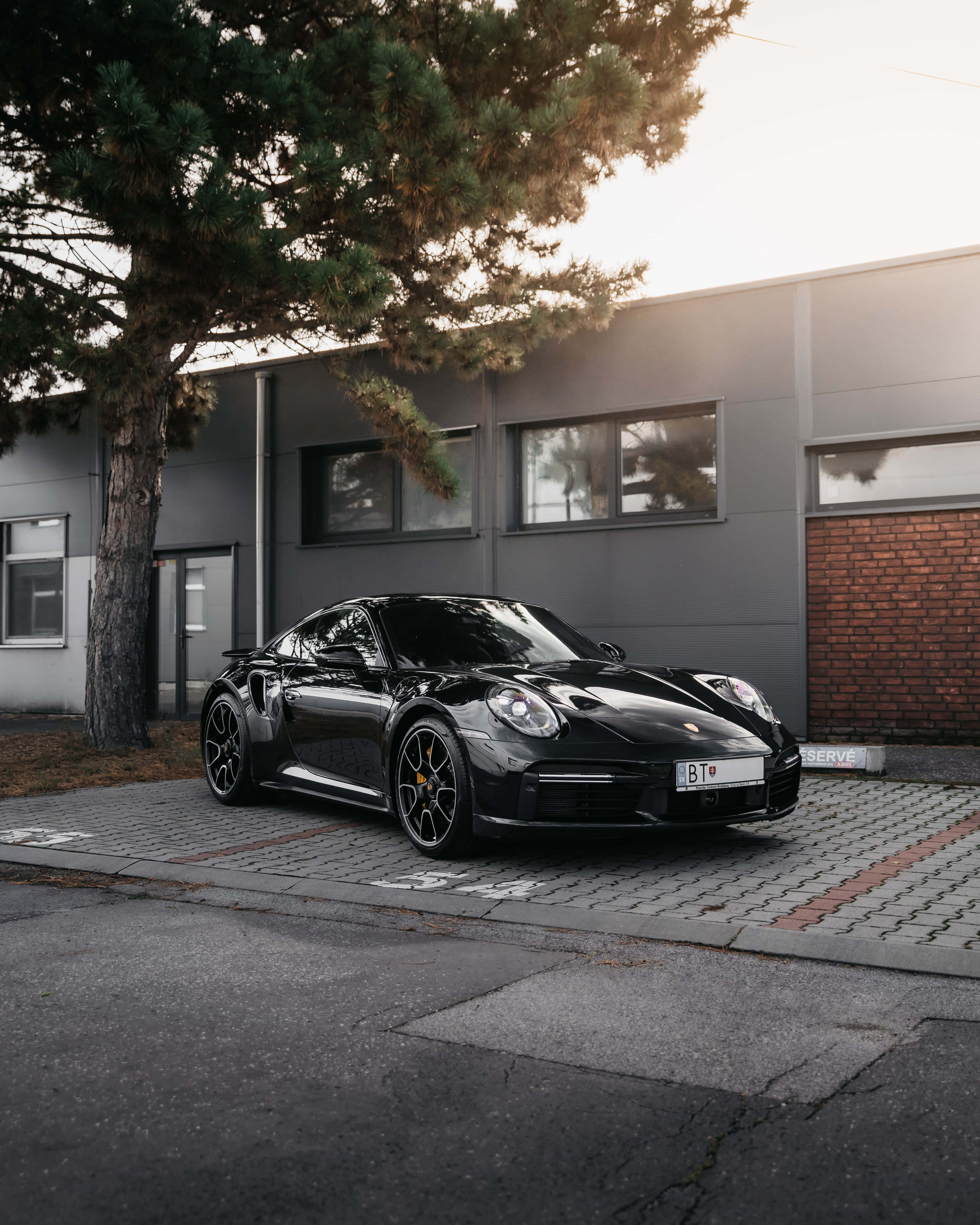 黑色 桌布 壁紙 — 一輛流線型黑色保時捷911 Turbo S停放在鋪砌的停車場，旁邊有樹木和現代建築，沐浴在柔和的金色時光中。