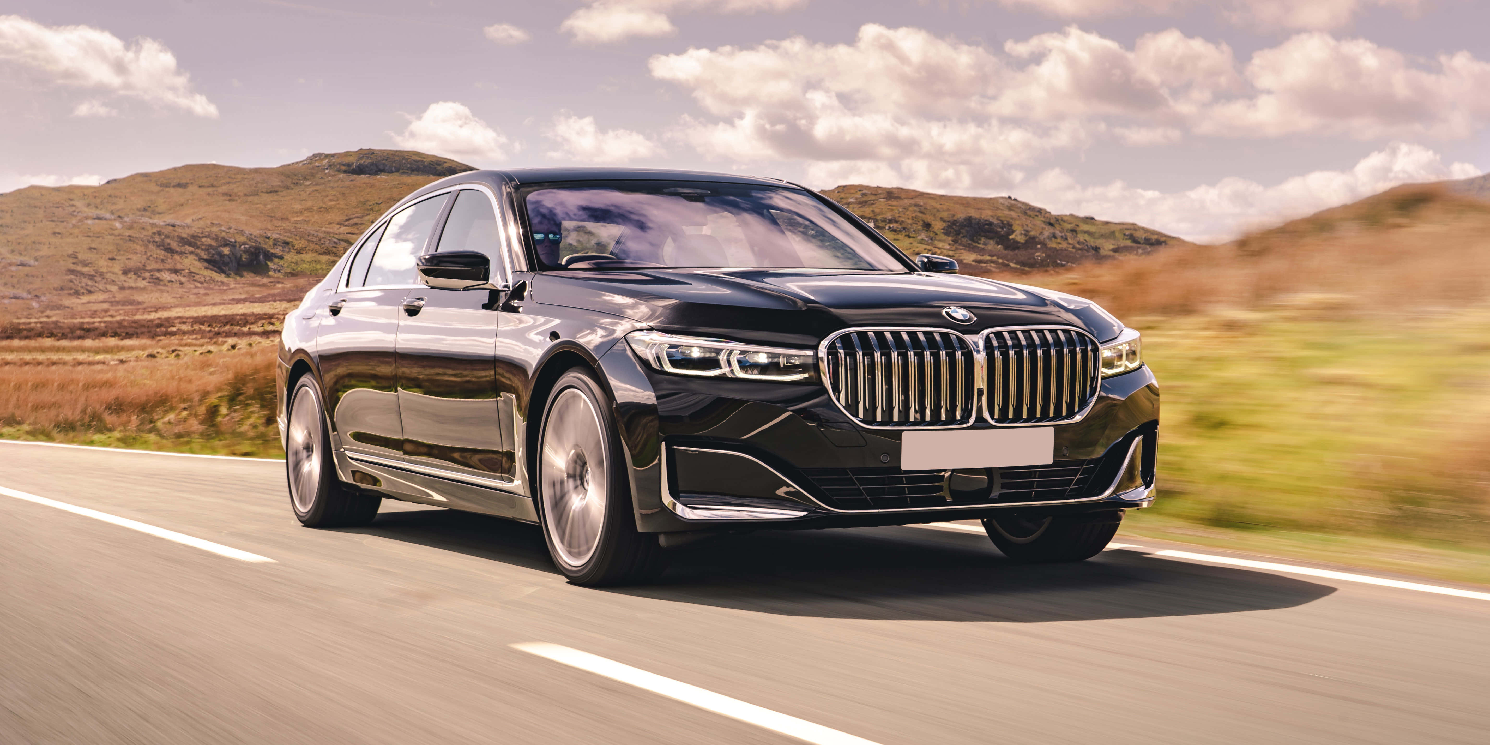 Bmw 7 Series 桌布 壁紙 — 一輛流線型黑色寶馬轎車在部分多雲的天空下，沿著蜿蜒的道路穿過起伏的金色山丘疾馳。
